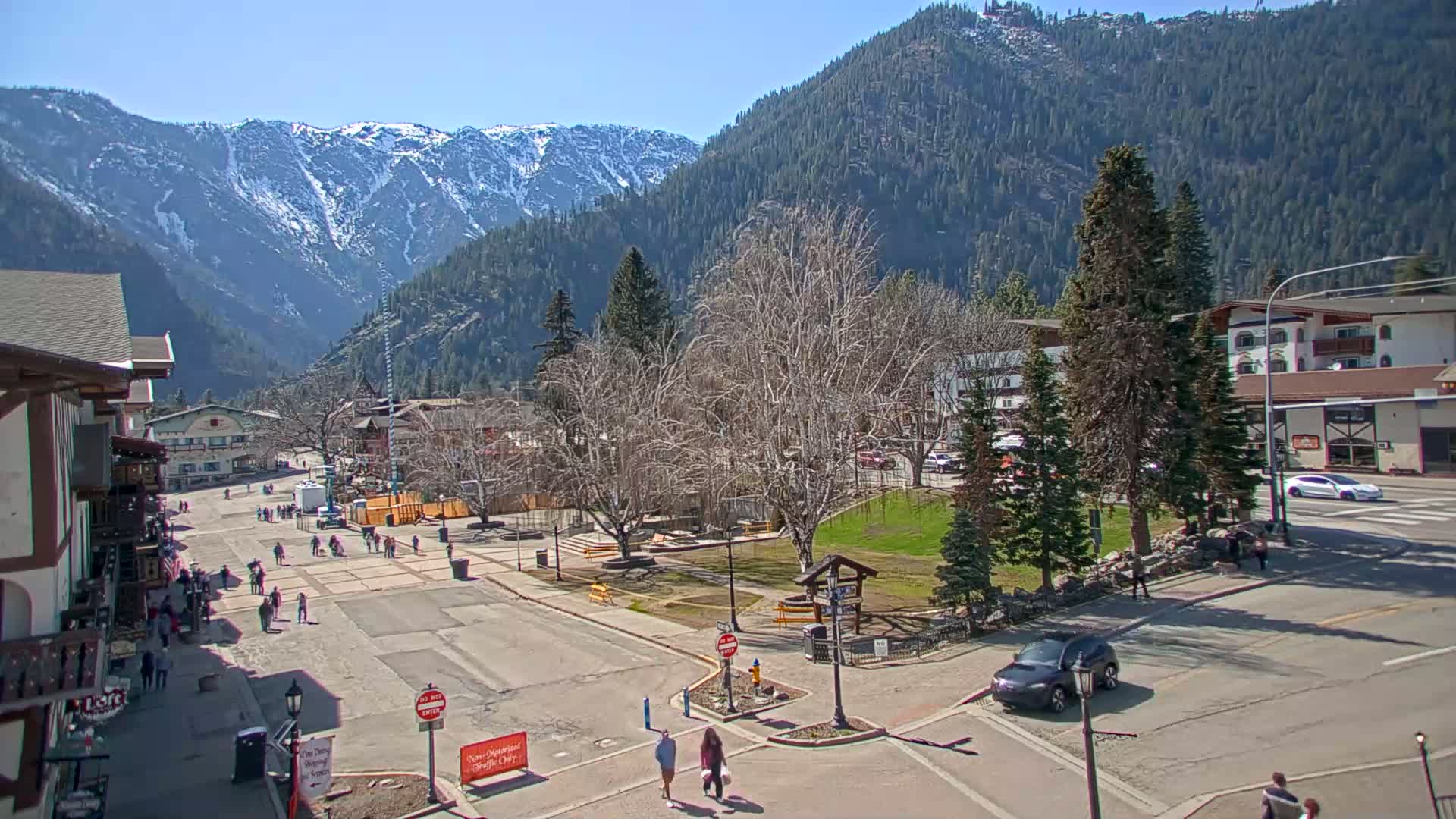 A sunny day in a mountain town shows a paved town square with pedestrians, cars, and buildings, all nestled against a backdrop of snow-capped mountains and evergreen trees.
