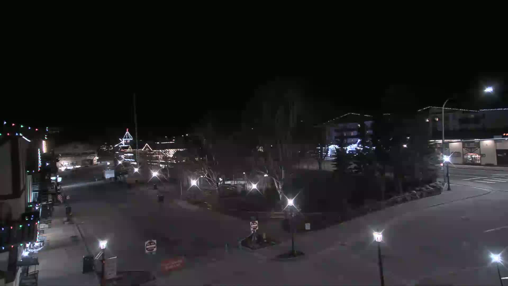 A nighttime view of a town square with buildings illuminated by streetlights and holiday decorations under a clear, dark sky.