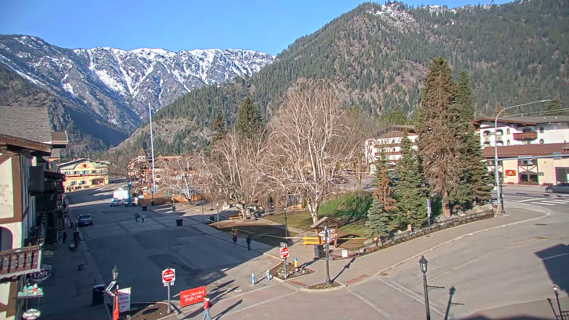 A mostly sunny day reveals a mountain town square with bare trees, snow-capped mountains in the background, and a few people walking around.