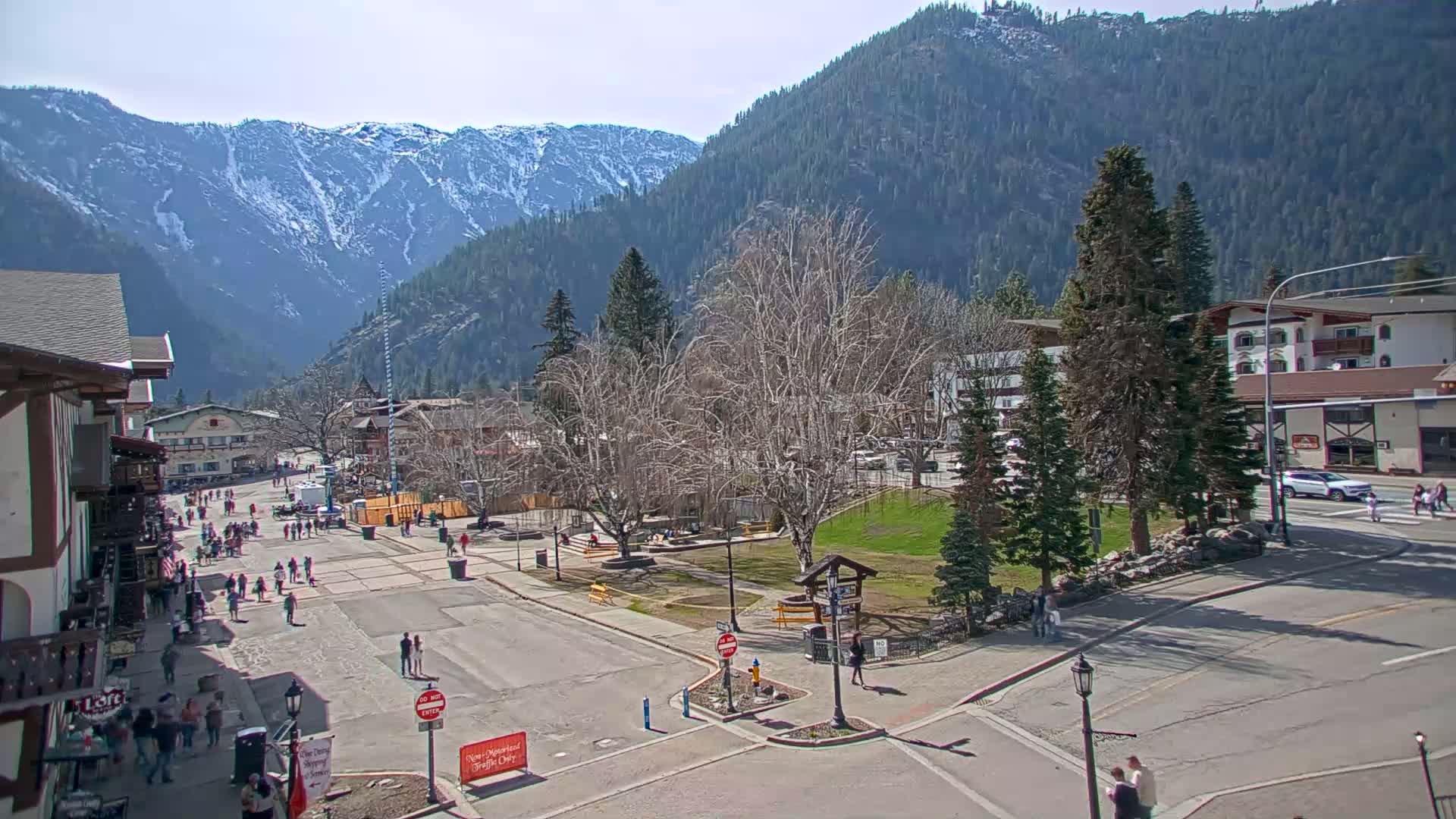 A sunny day in a mountain town shows a pedestrian area with people walking near buildings, a small park, and snow-capped mountains in the background.
