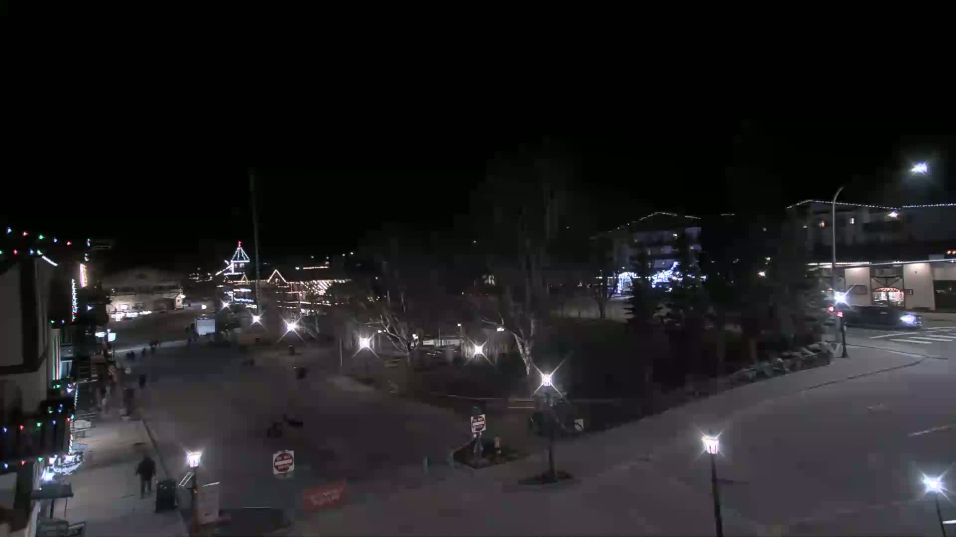 A nighttime view of a town square with buildings decorated with lights, a few pedestrians, and cars driving on a street.
