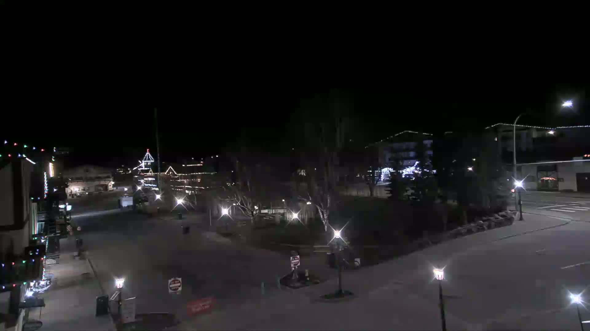 A nighttime, mostly empty town square with several buildings decorated with Christmas lights.