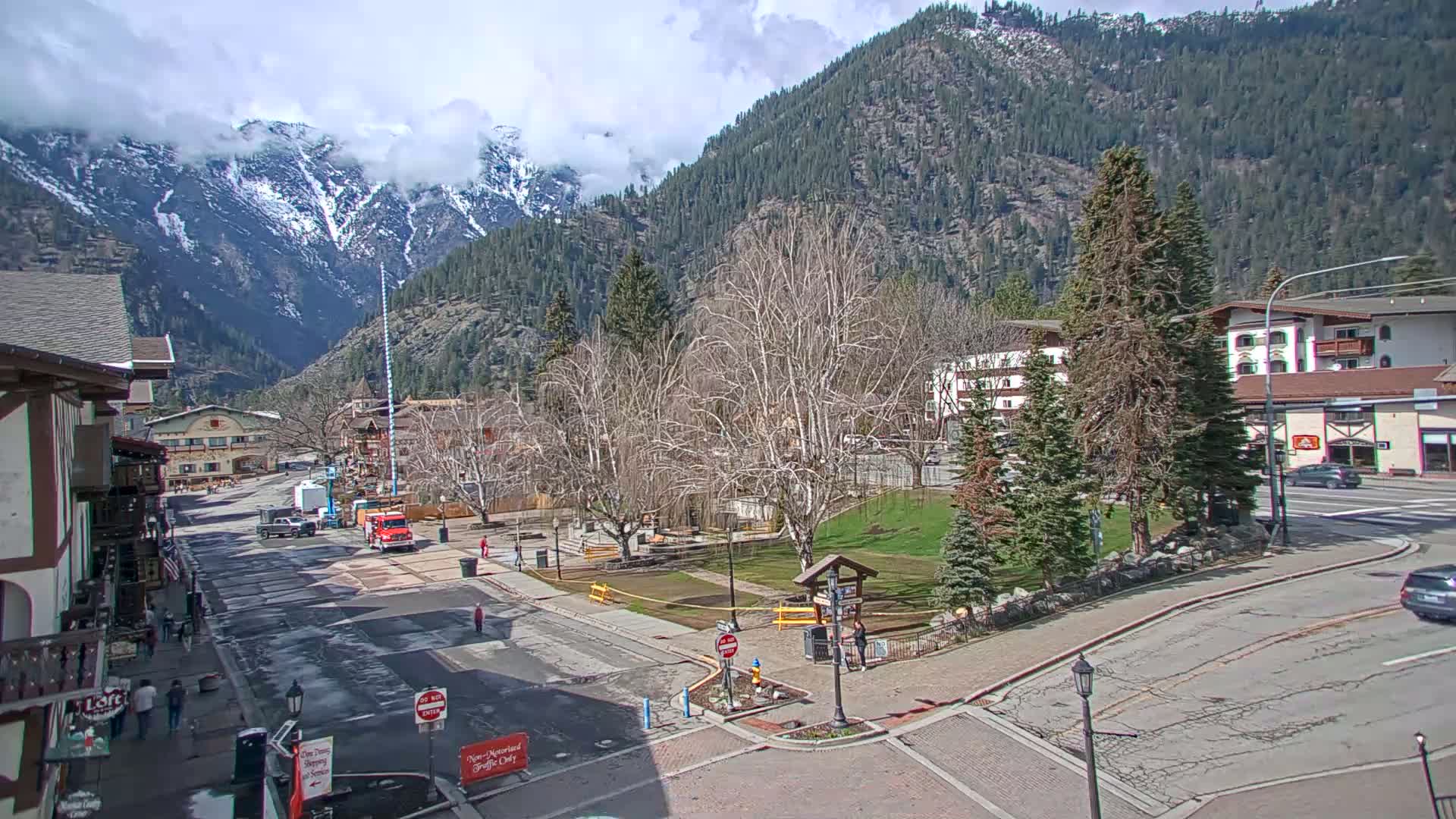 A partly cloudy day reveals a mountain town nestled at the base of snow-capped mountains, with a mostly empty street and a small park visible in the foreground.
