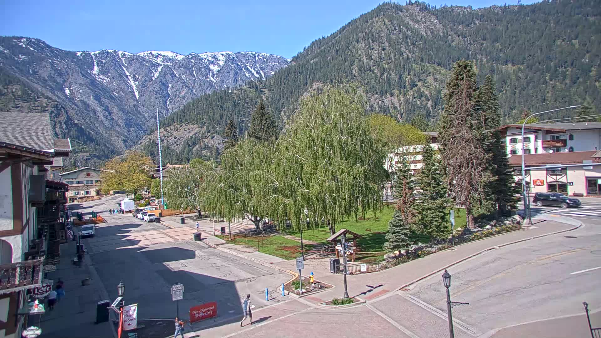 A sunny view of a town square nestled in a valley, with snow-capped mountains in the background.