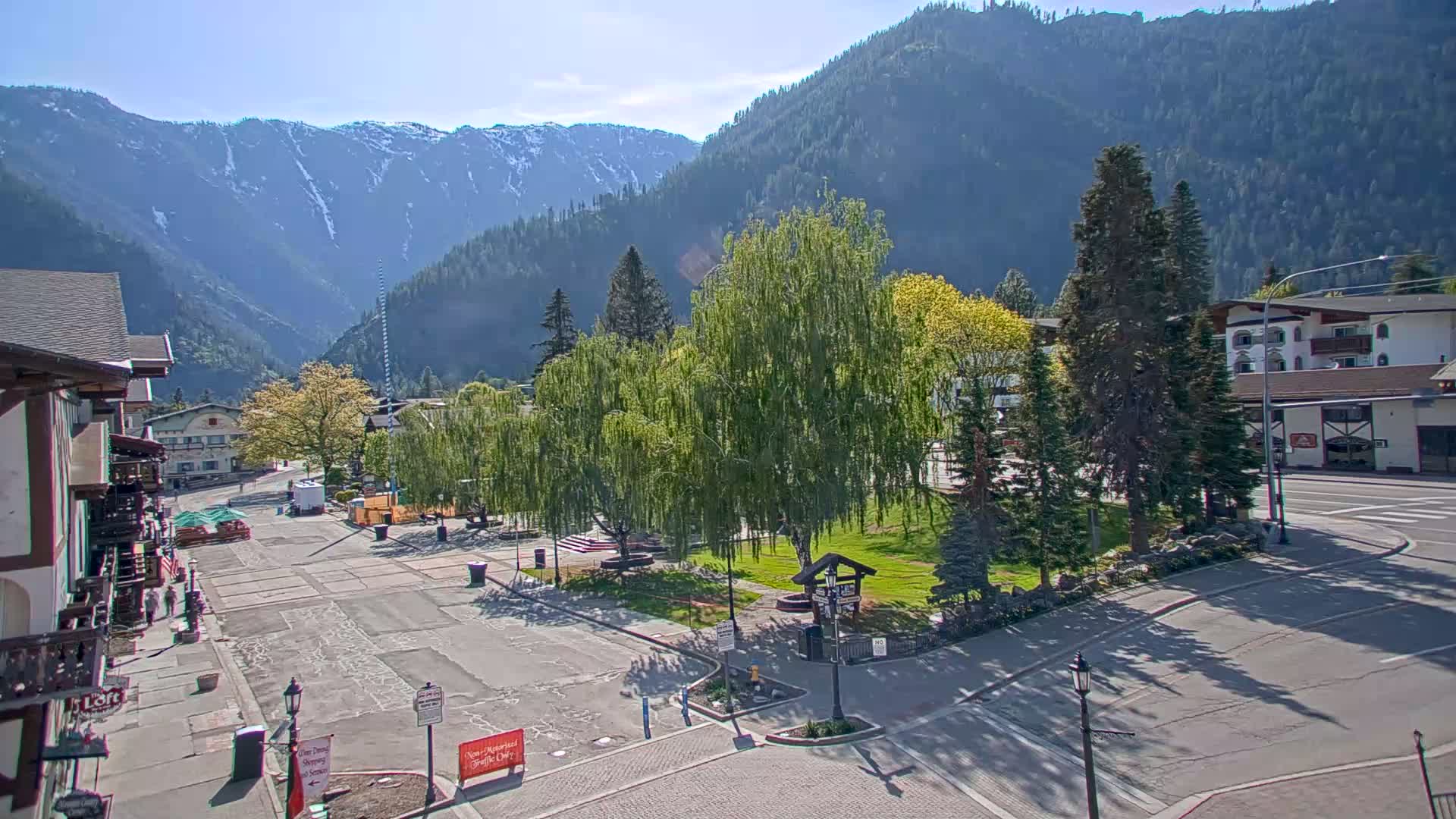 A mostly empty town square, with green trees and a snow-capped mountain range in the background, under sunny skies.