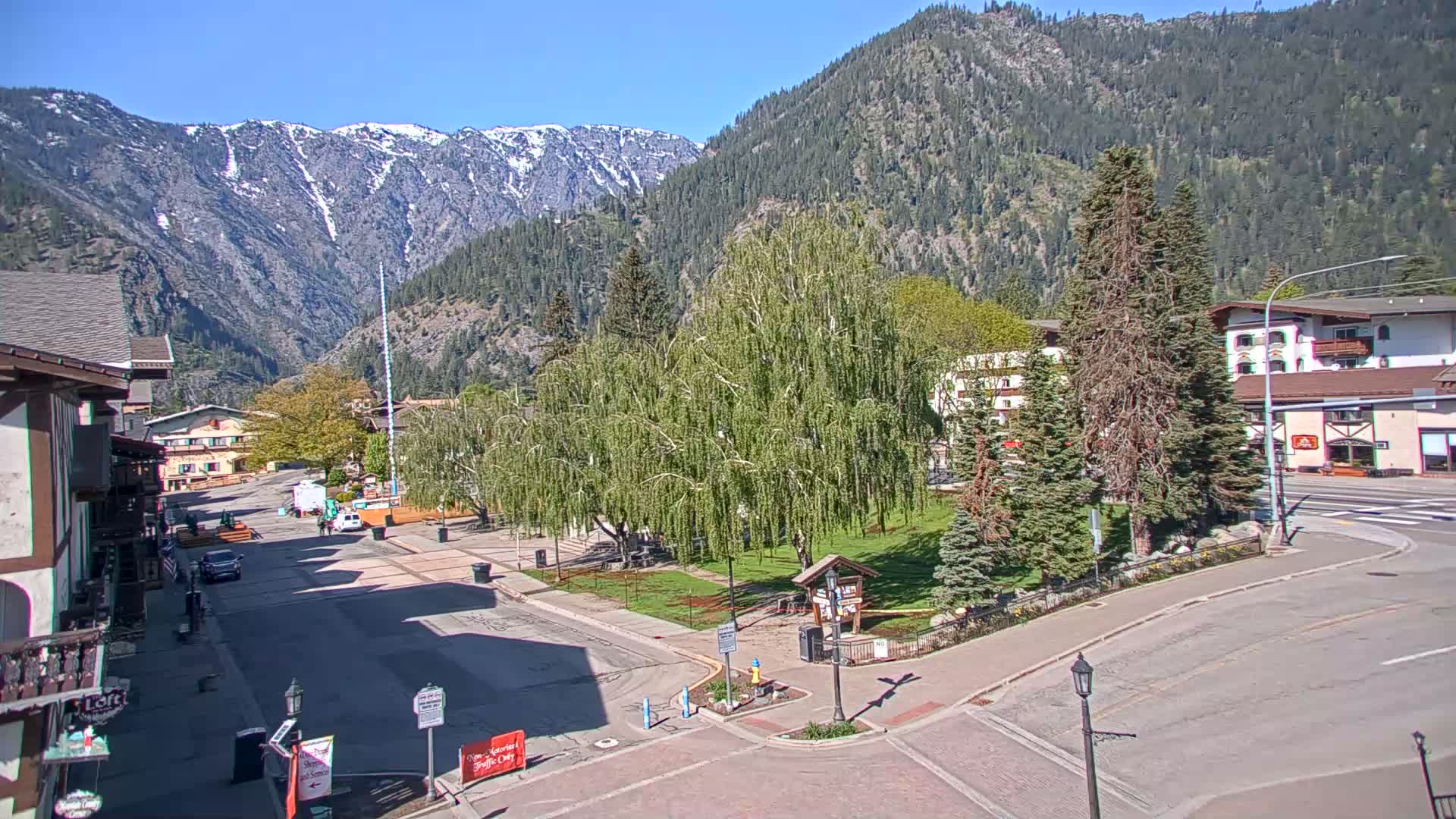 A sunny view of a town square nestled in a valley, surrounded by mountains with patches of snow, and leafy green trees.