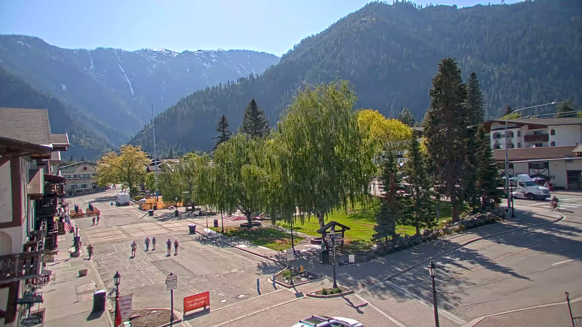 A mostly empty town square with a few pedestrians, surrounded by buildings and trees, sits nestled in a valley beneath snow-capped mountains on a sunny day.