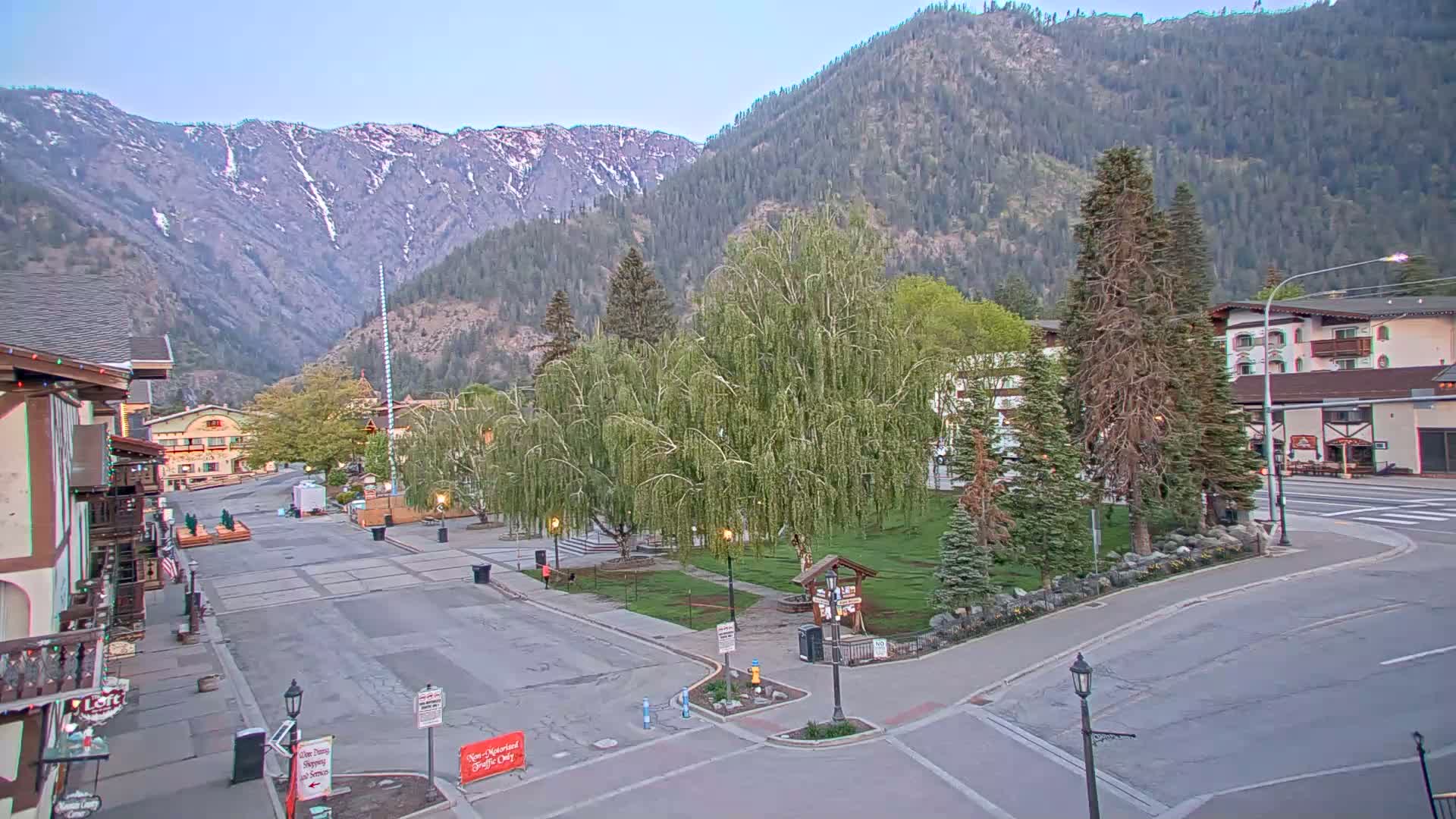 A mostly empty town square with buildings and trees, nestled in a valley beneath snow-capped mountains on a clear day.