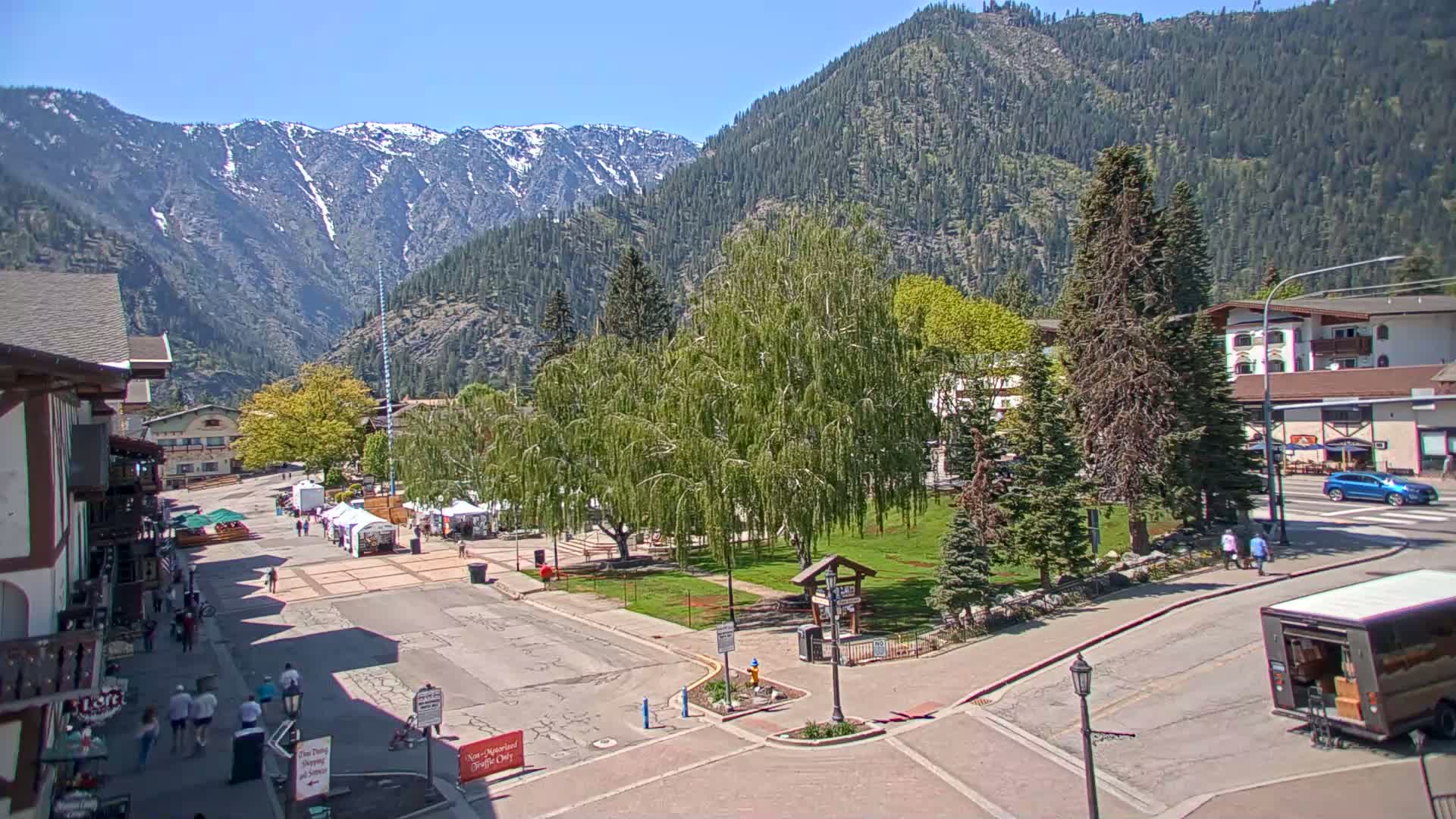 A town square with vendor booths and pedestrians is nestled in a valley between snow-capped mountains under a clear blue sky.