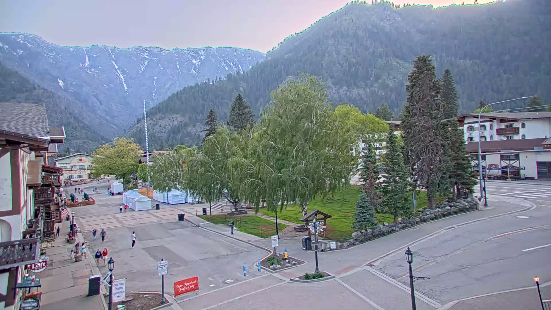 A mostly empty town square, with a few pedestrians, sits between buildings and a large, snow-capped mountain range under a partly cloudy sky.