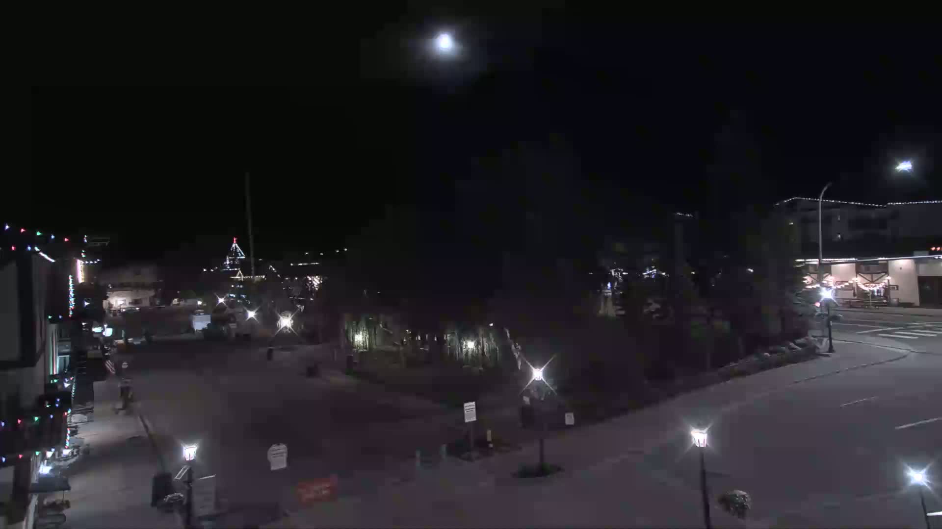 A mostly empty, nighttime street scene shows some buildings with Christmas lights, trees, and streetlights under a bright moon.