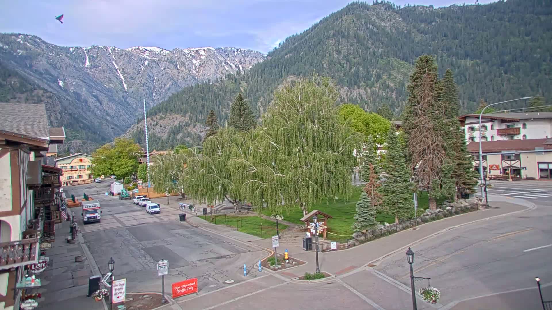 A partly sunny day reveals a mountain town square with a small park, buildings, and cars, all nestled in a valley against a backdrop of snow-capped mountains.