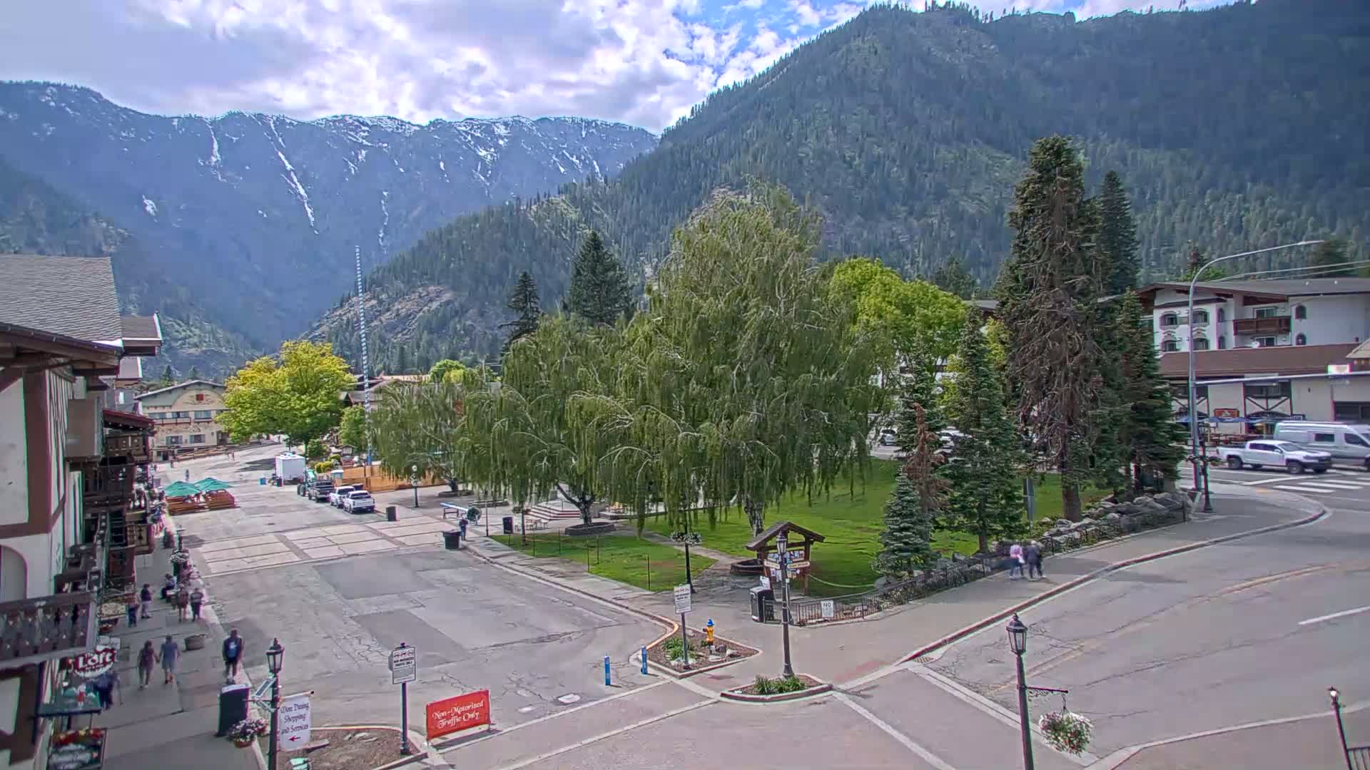 A partly cloudy day reveals a mountain town square with a small park, surrounded by buildings and a few people walking, all set against a backdrop of snow-capped mountains.