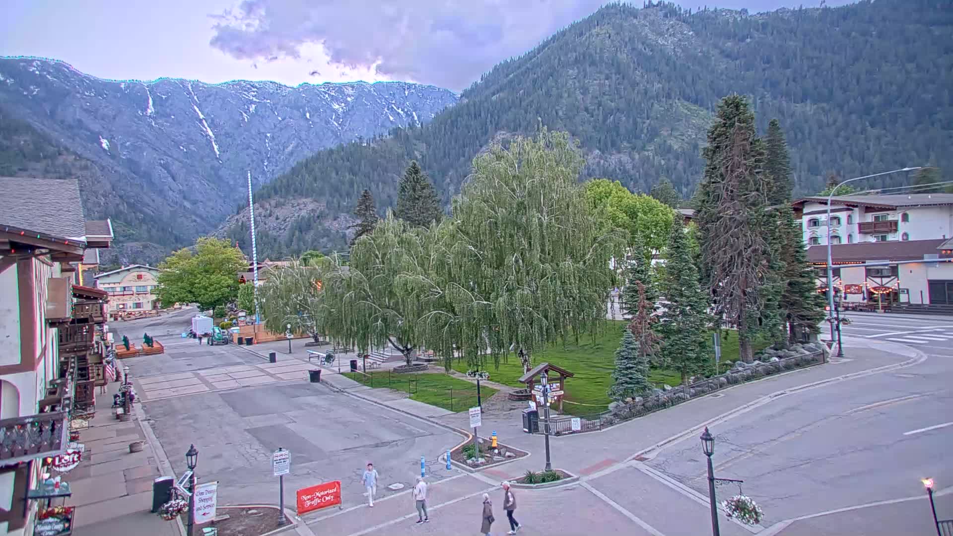 A mostly sunny town square nestled in a valley between snow-capped mountains, with several people walking around.