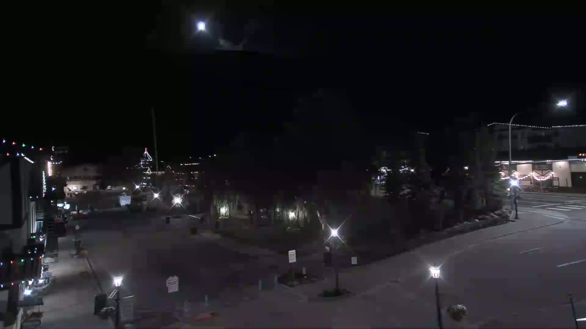 A moonlit night scene shows a mostly empty town square with some buildings, streetlights, and sparsely-placed trees.