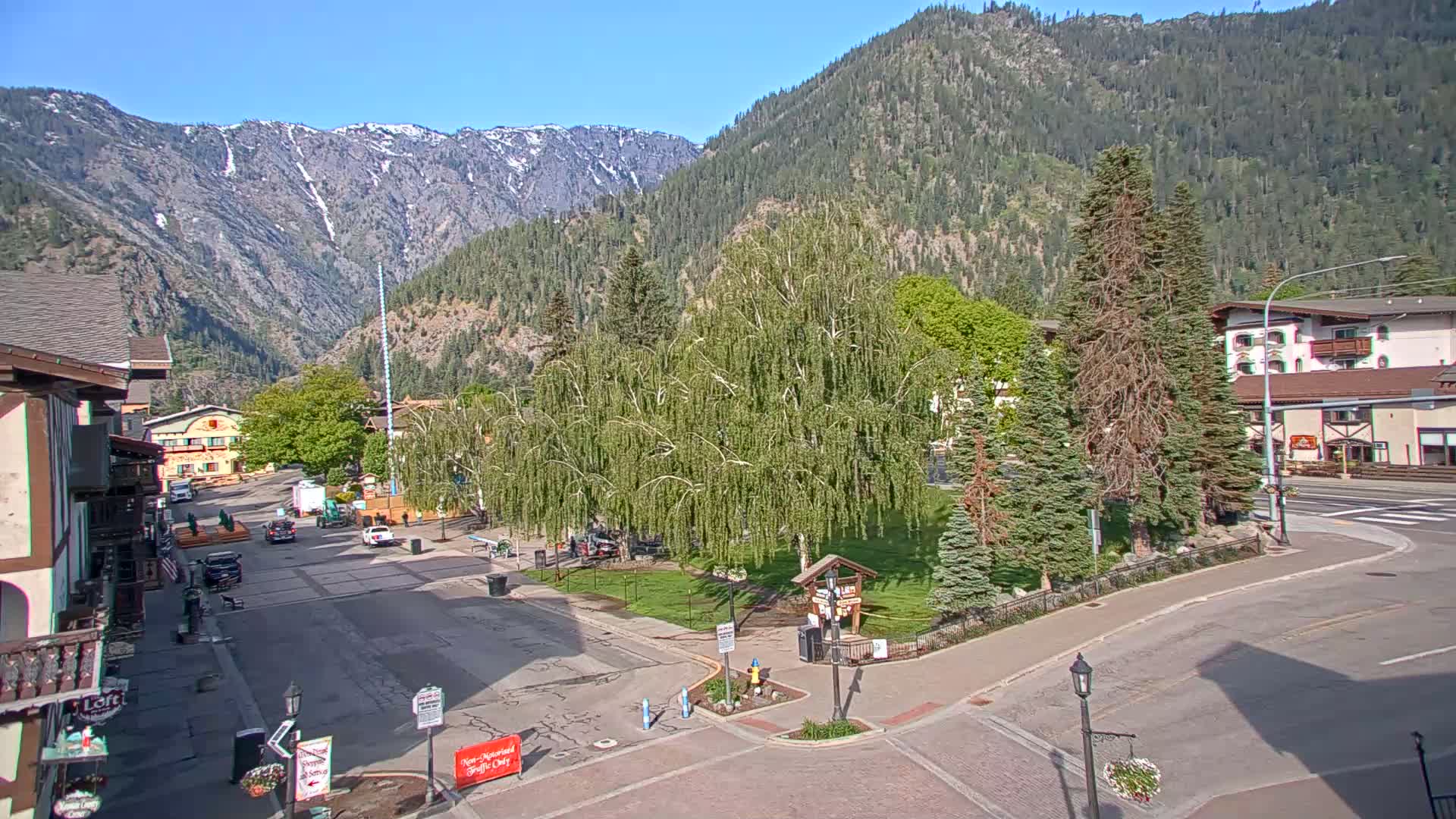 A town square with a grassy area and several trees is nestled between buildings and mountains on a sunny day.