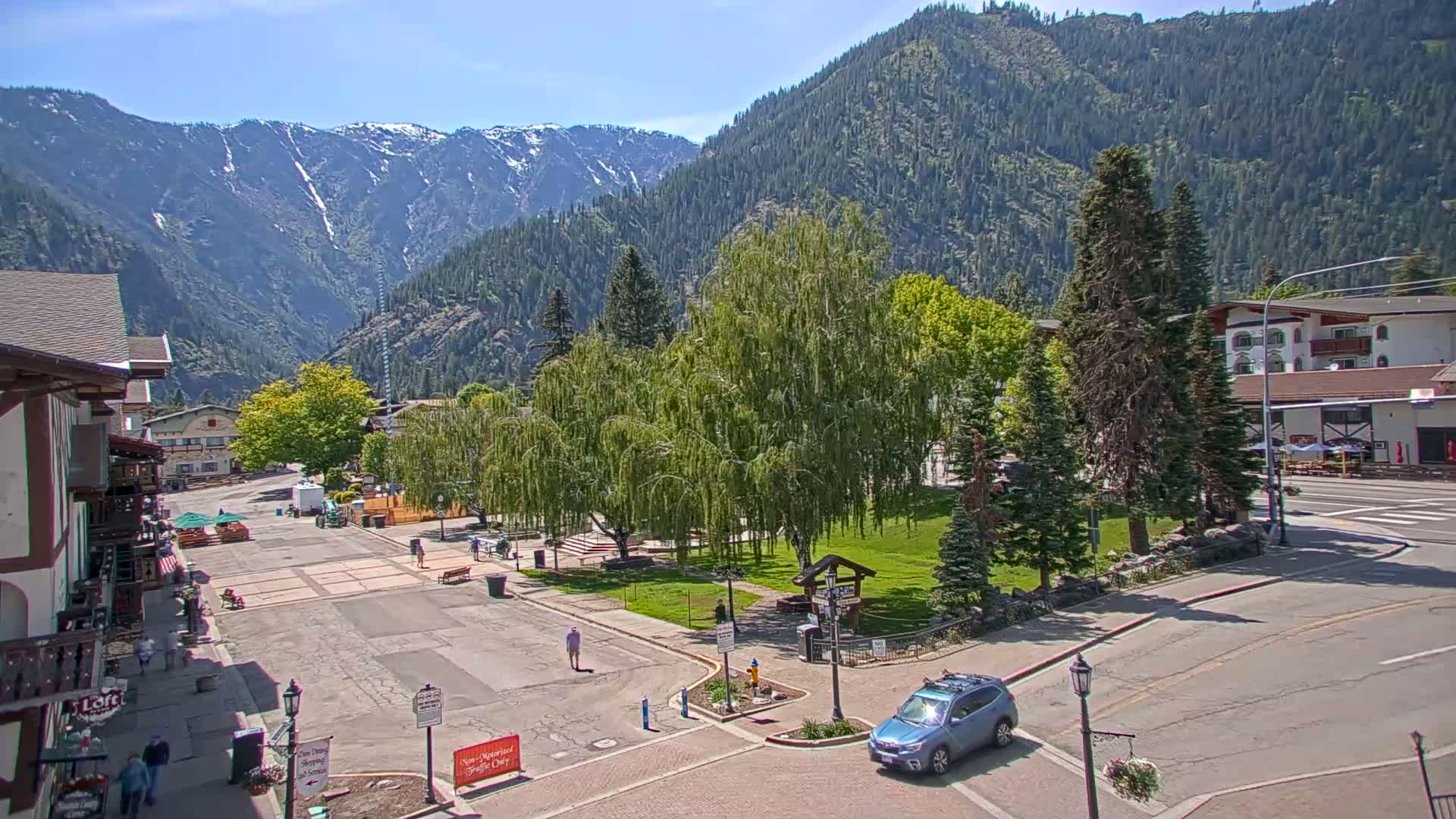 A sunny mountain town square with a few people, some cars, and green trees, backed by snow-capped mountains.