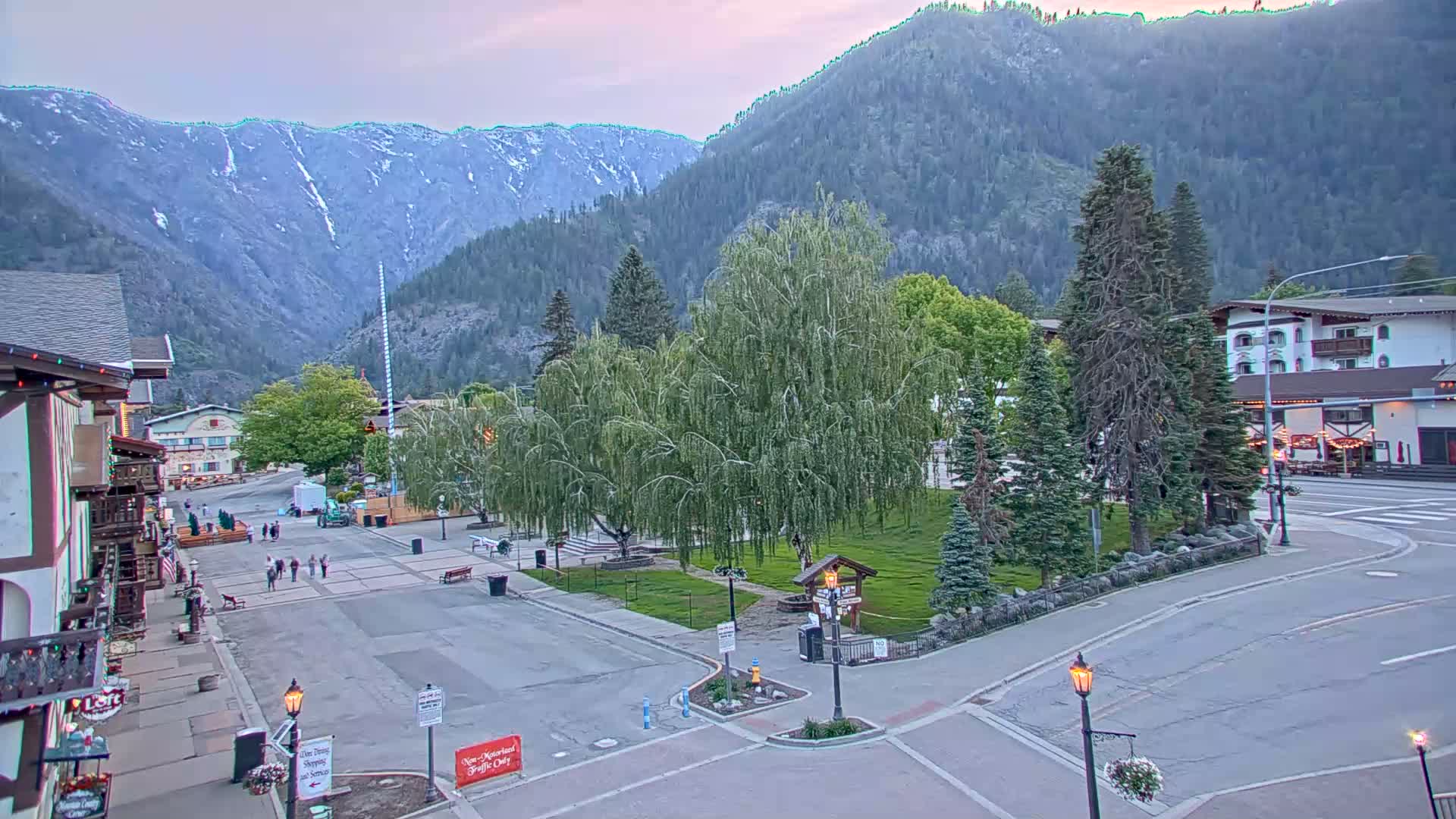 A mountain town square with a weeping willow tree in the center, under a partly cloudy evening sky, is surrounded by buildings and a few pedestrians.