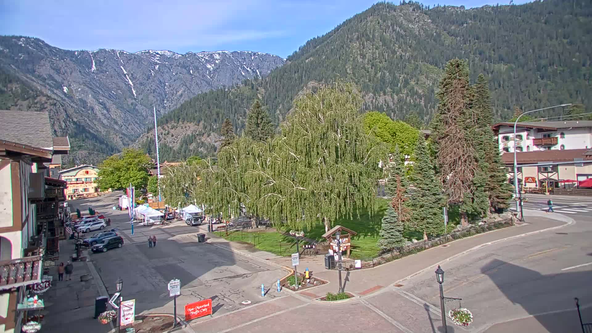A mountain town square with a grassy park, several buildings, and cars parked along the streets, under a clear sunny sky, with mountains in the background showing some snow.