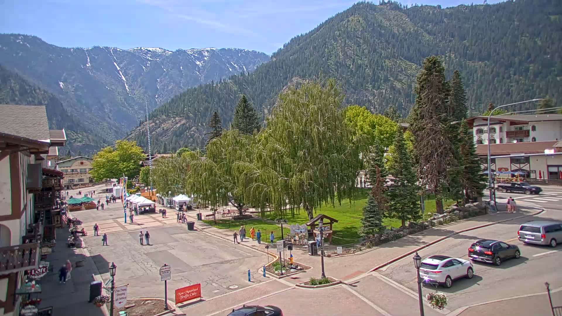 A town square with a grassy area and several trees is surrounded by buildings, with snow-capped mountains in the background under a sunny sky.