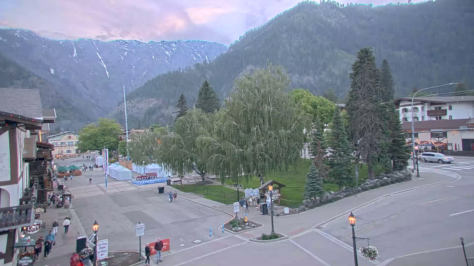 A town square with people walking around, surrounded by buildings and trees, sits at the base of a mountain range under a partly cloudy sky.