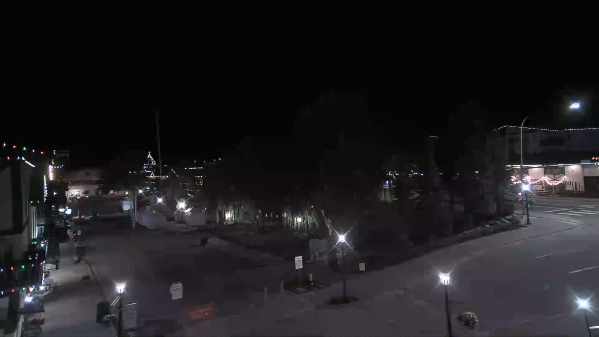 A nighttime view of a town square with sparsely lit buildings and trees, under a clear, dark sky.