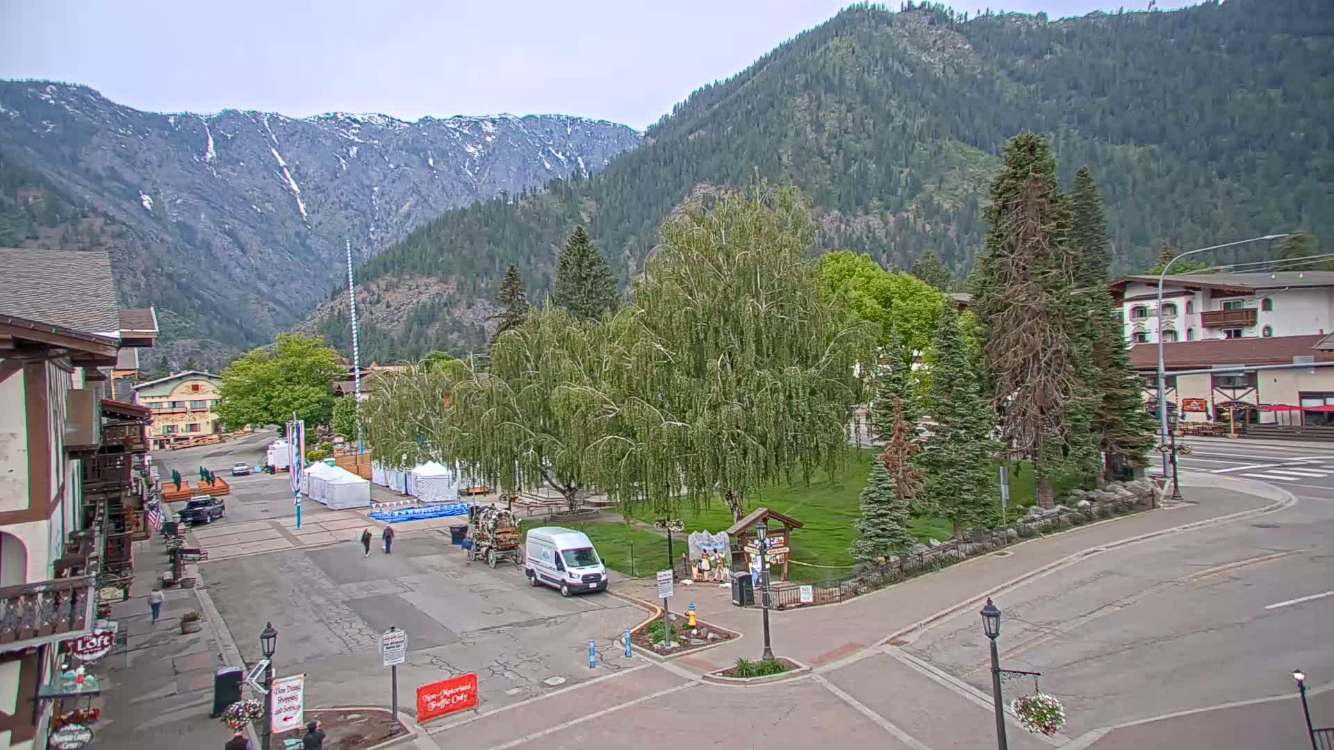 A town square with a few people and vehicles, surrounded by buildings and trees, sits against a backdrop of mountains under an overcast sky.