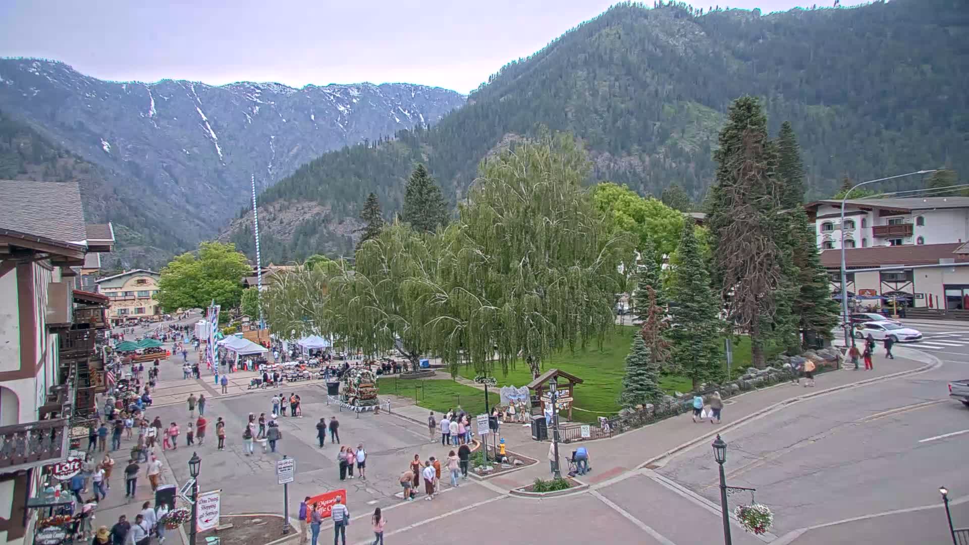 A town square filled with people, surrounded by buildings and trees, nestled in a valley between snow-capped mountains under an overcast sky.