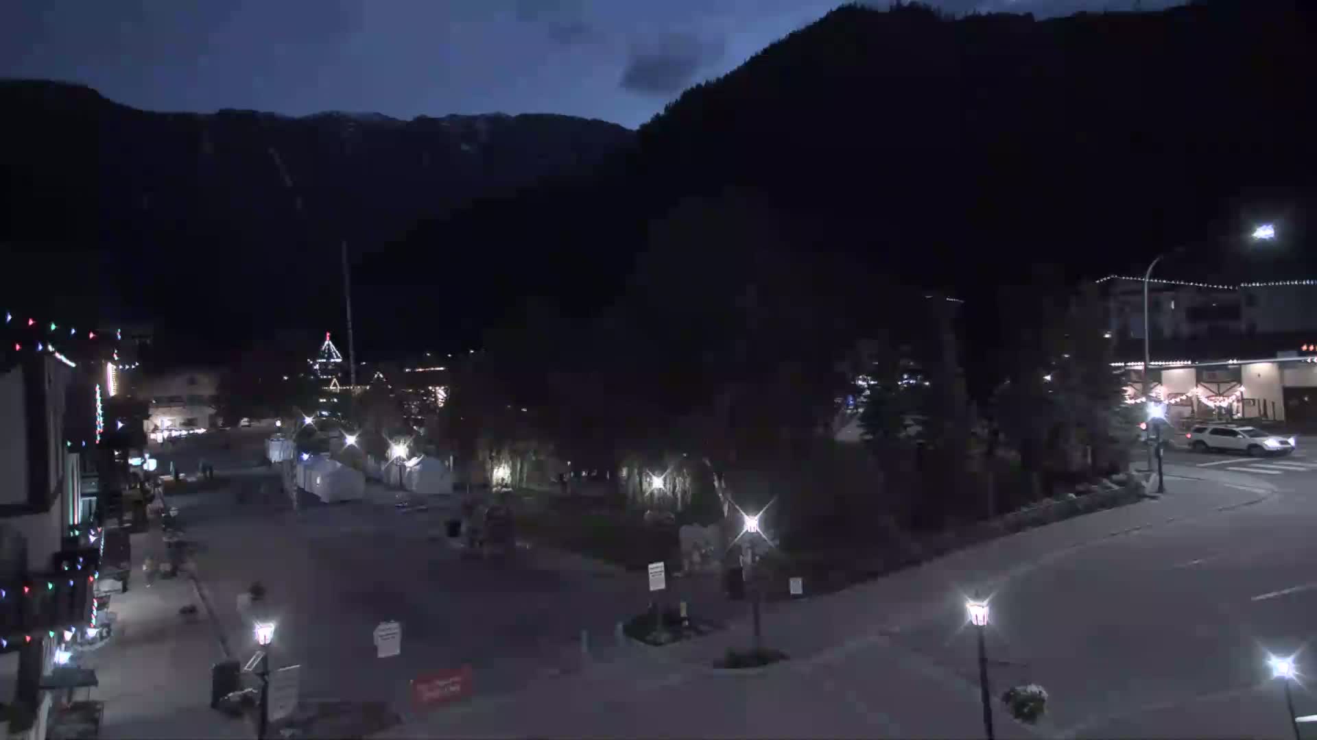 A nighttime view of a town square, nestled against a dark mountainside under a mostly clear, dark sky, with a few cars on the roads and festive lights illuminating buildings.