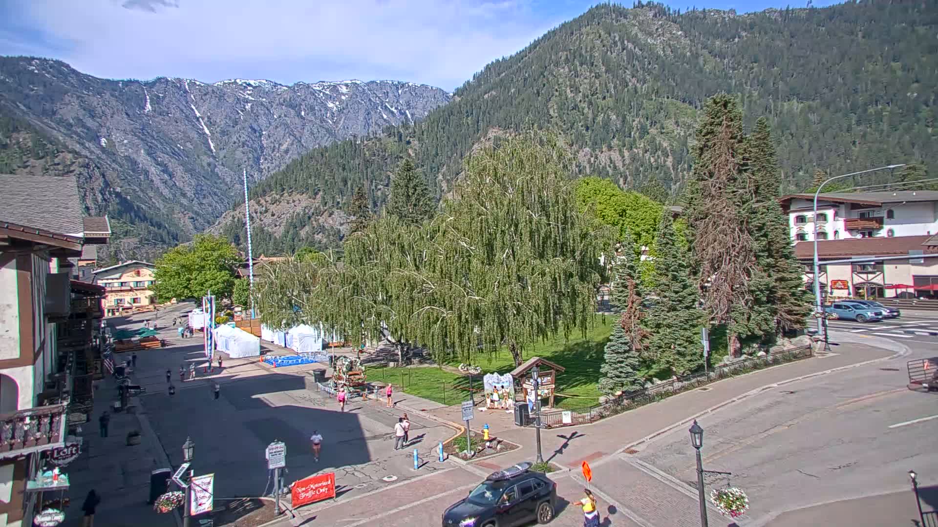 A sunny town square nestled in a valley between snow-capped mountains, with people walking around and cars parked on the streets.