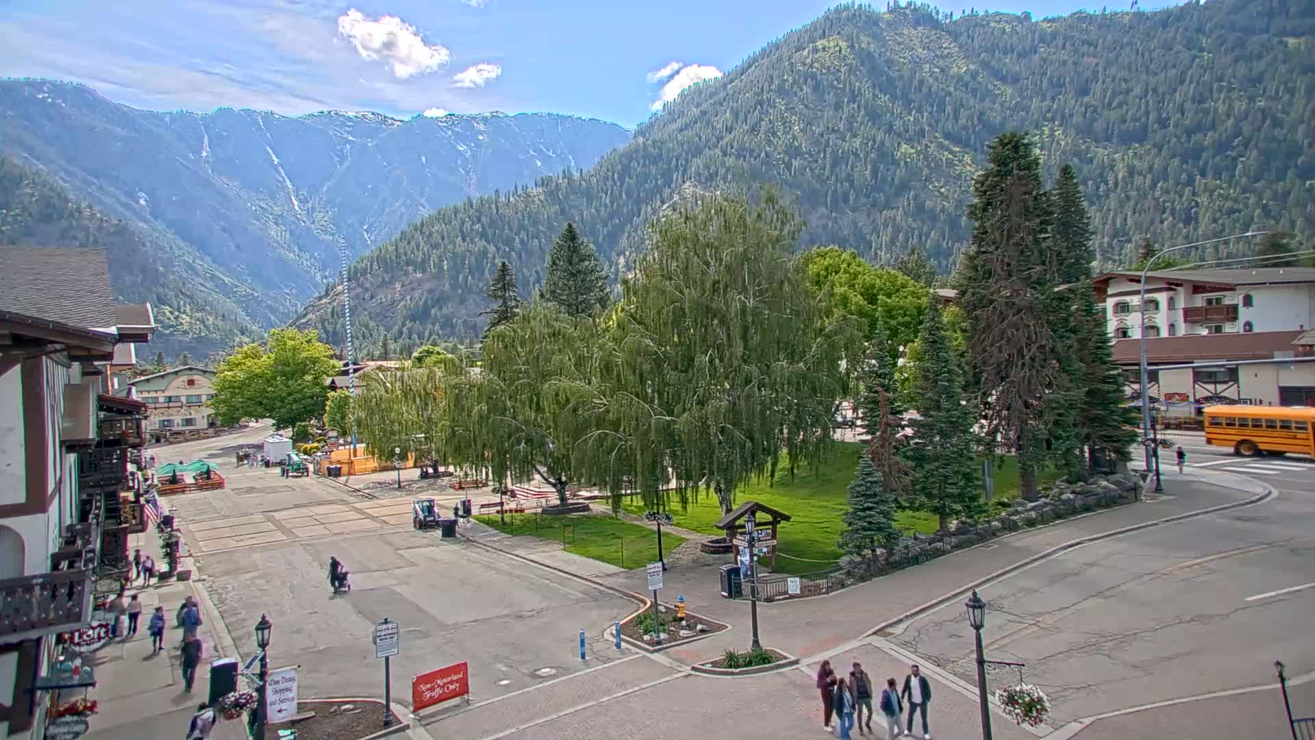 A mountain town square with a grassy park and several buildings, under a mostly sunny sky, is viewed from above.