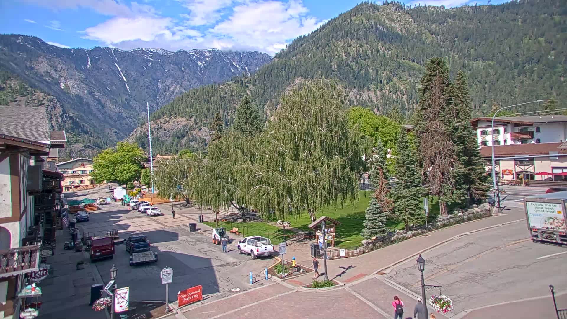 A town square with a weeping willow tree and surrounding buildings is nestled in a valley under a partly cloudy sky, with snow-capped mountains in the background.