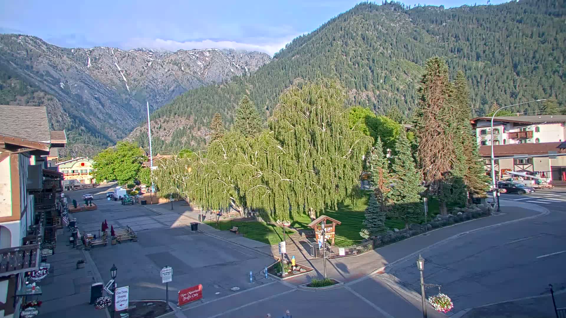 A mountain town square with a small park in the center, surrounded by buildings and trees, under a mostly clear sky with some clouds and mountains in the background.