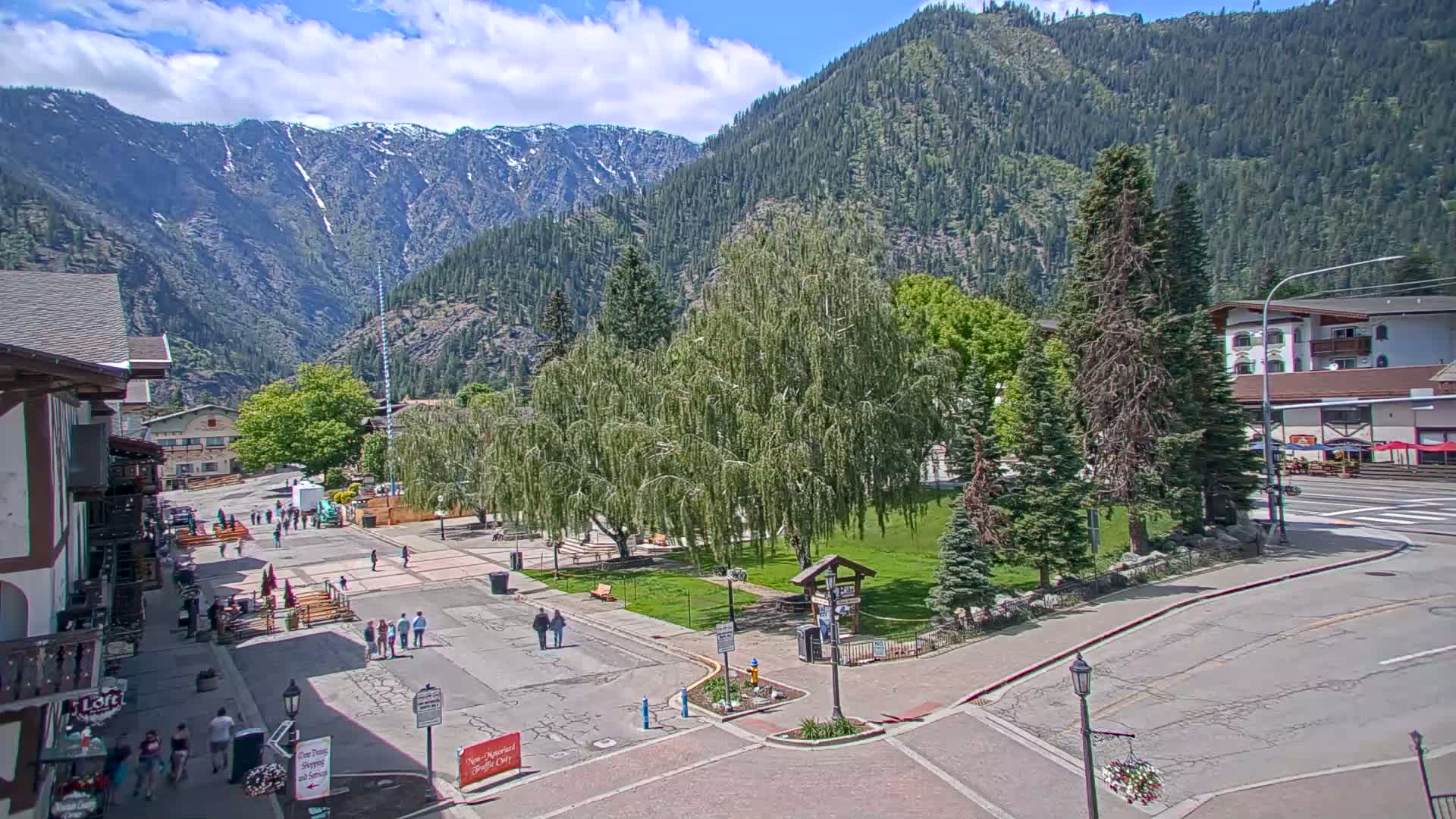 A mountain town square with a grassy park, several buildings, and people walking around on a sunny day with partly cloudy skies and snow-capped mountains in the background.