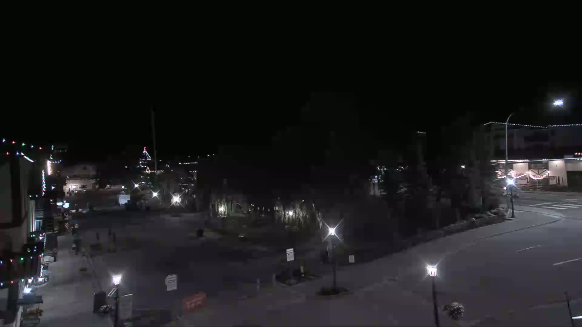A nighttime, mostly empty town square is illuminated by streetlights, with a few pedestrians visible and some buildings decorated with lights.