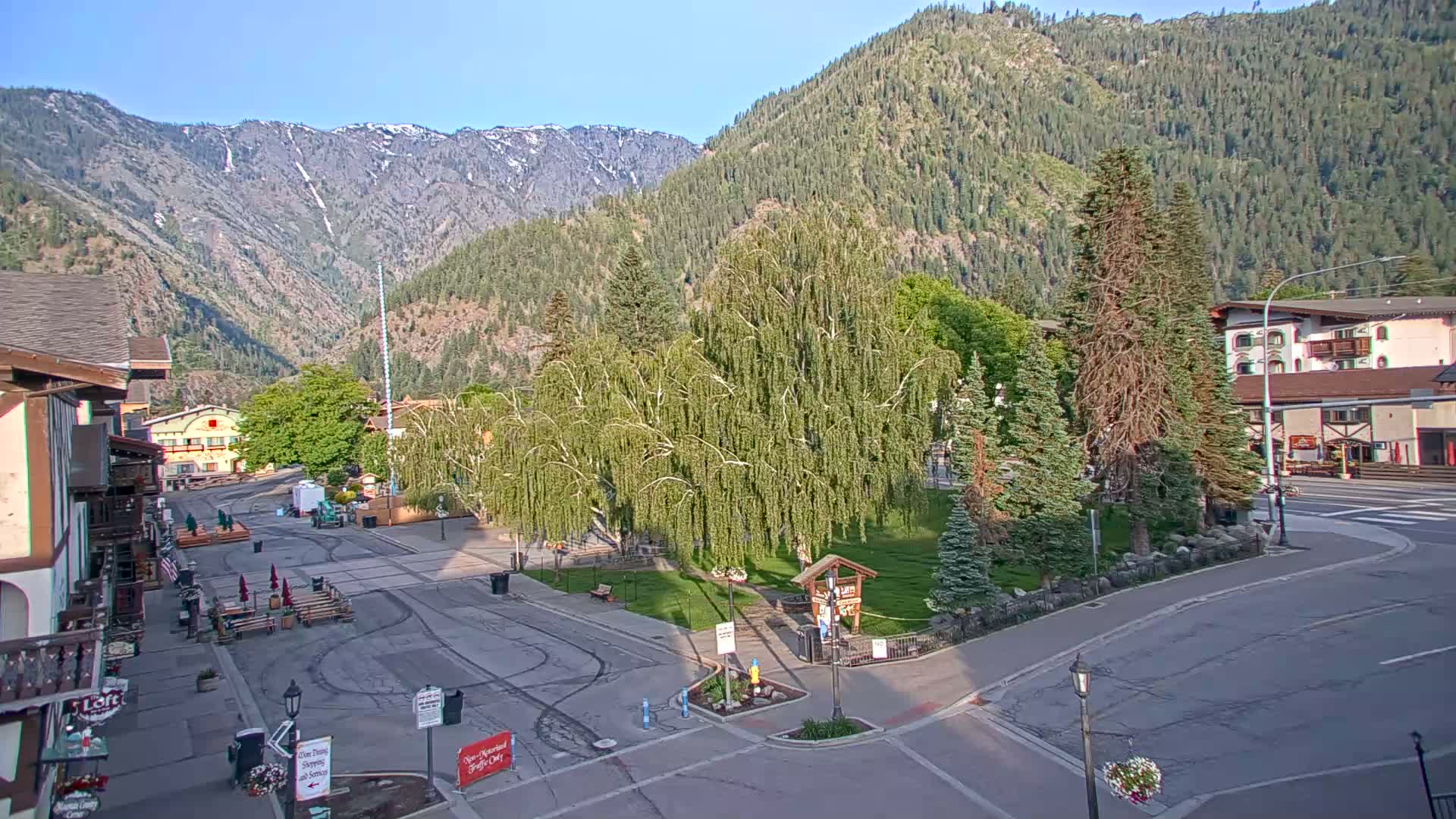 A town square with buildings and trees sits in a valley, nestled between mountains under a clear, sunny sky.