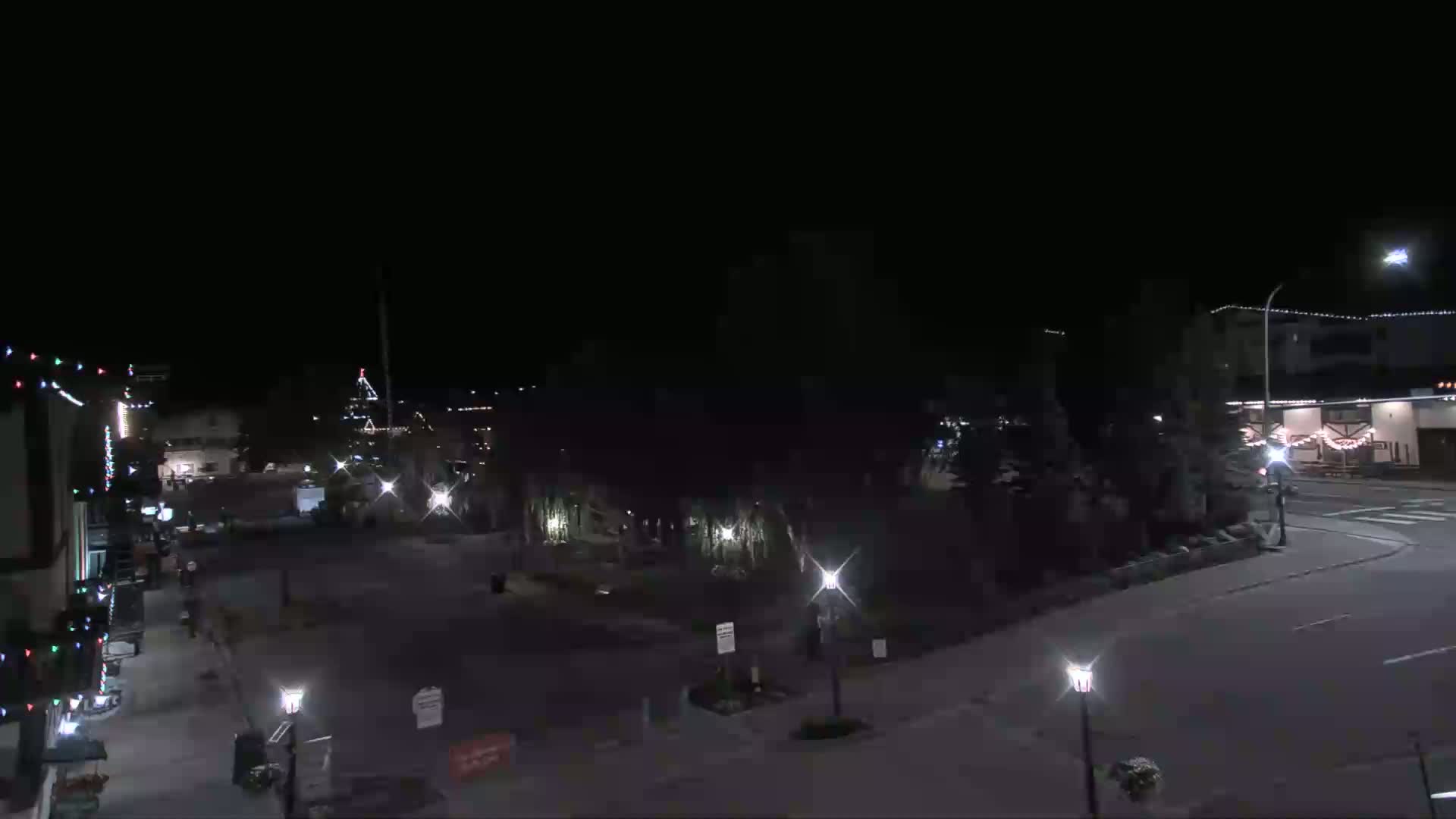 A nighttime, street-level view of a sparsely populated town square with some buildings decorated with lights.