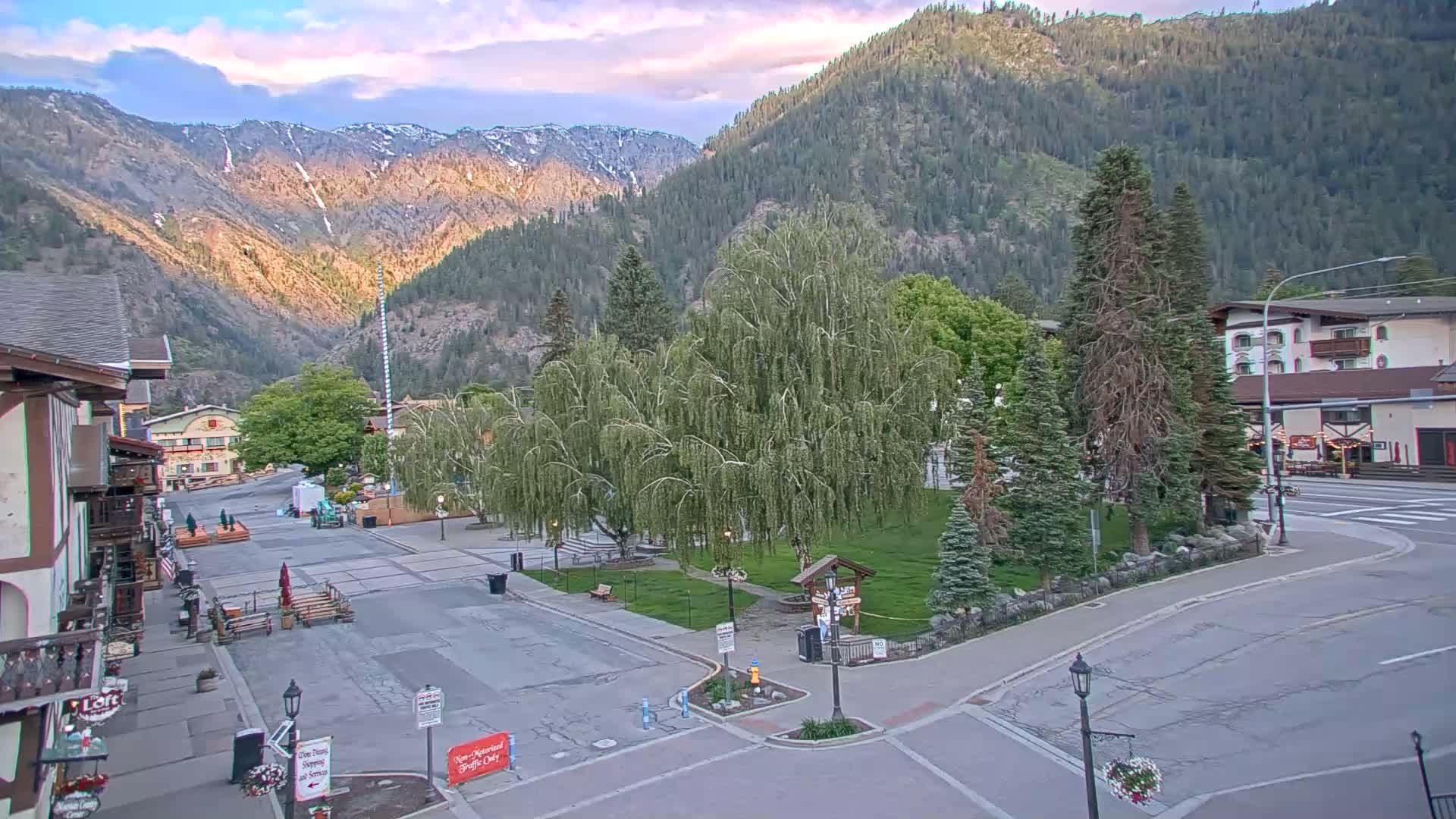 A mostly empty town square sits nestled between buildings and mountains under a partly cloudy sky.