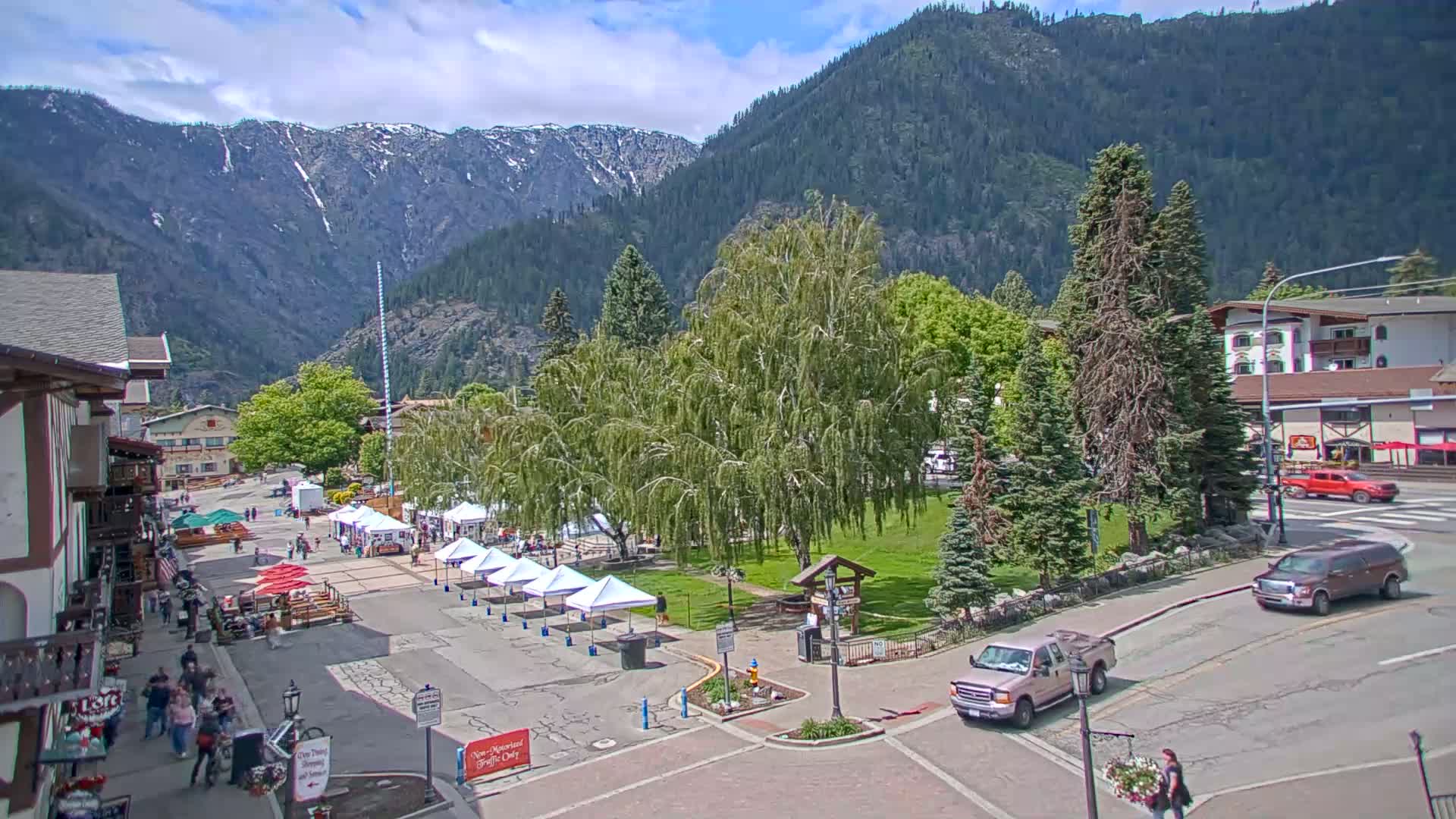 A town square with several market stalls is nestled in a valley between snow-capped mountains on a partly sunny day.