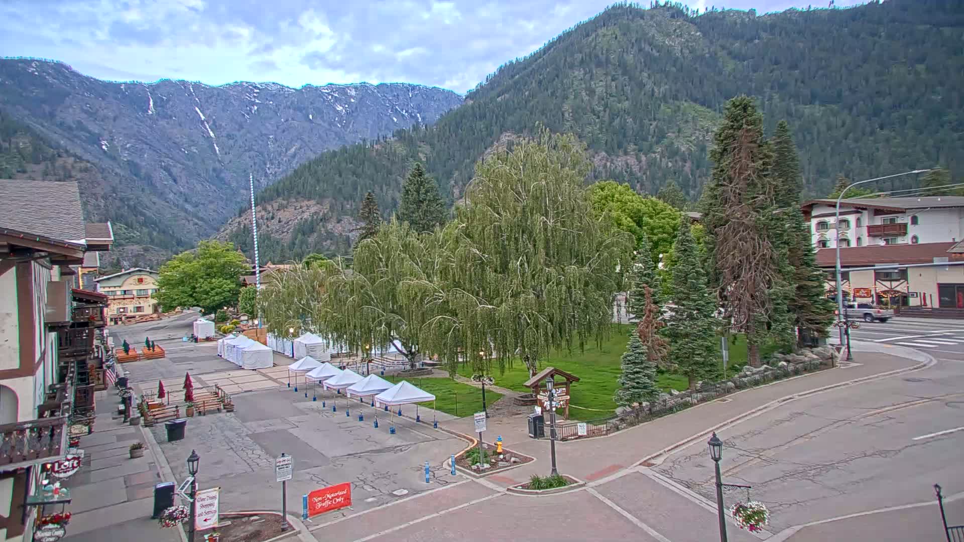 A mostly sunny day reveals a town square with several white tents, a large weeping willow tree, and buildings nestled at the base of a mountain range under a partly cloudy sky.