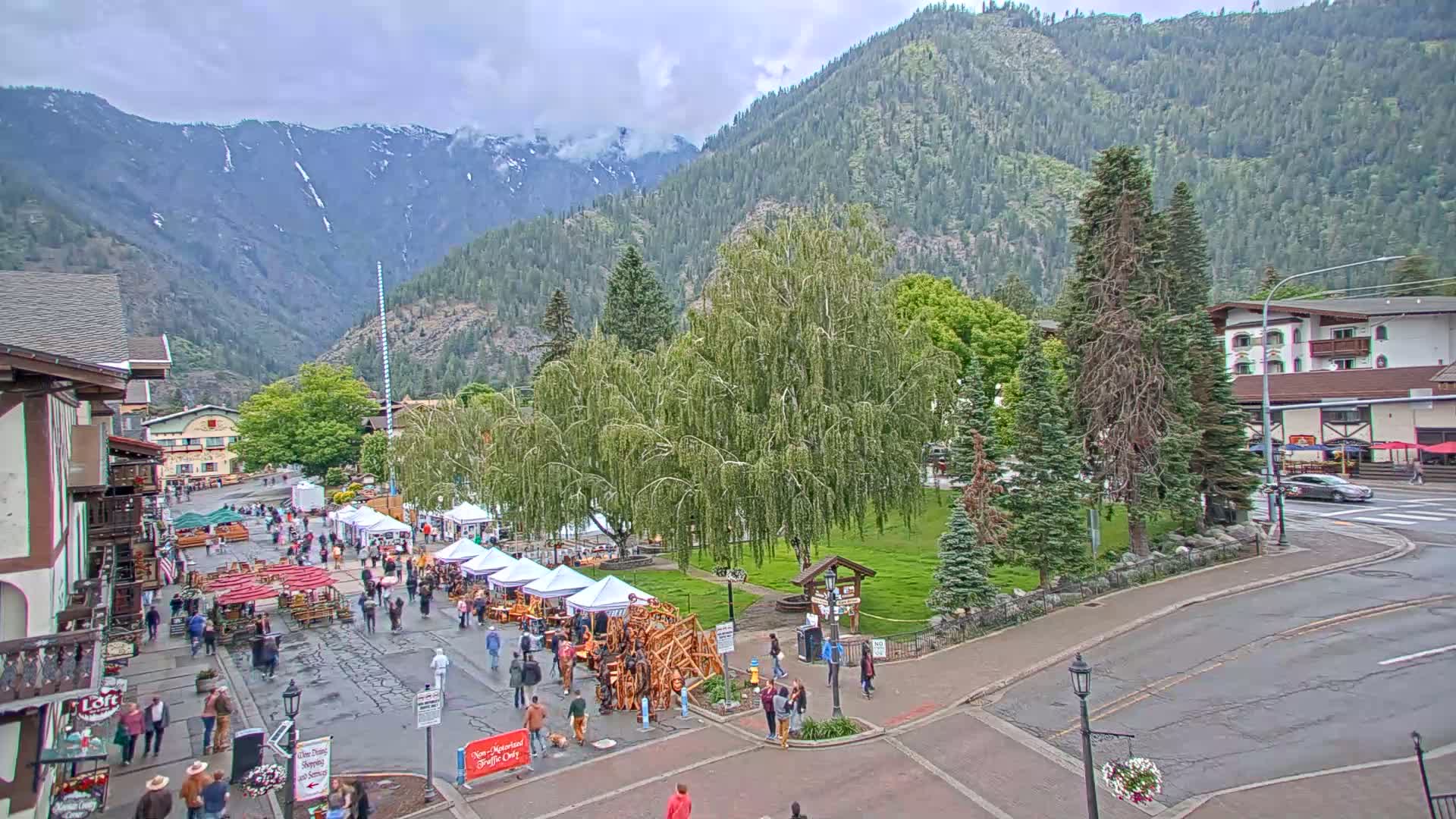 A town square with numerous market stalls and people walking around, situated in a valley with overcast skies and snow-capped mountains in the background.