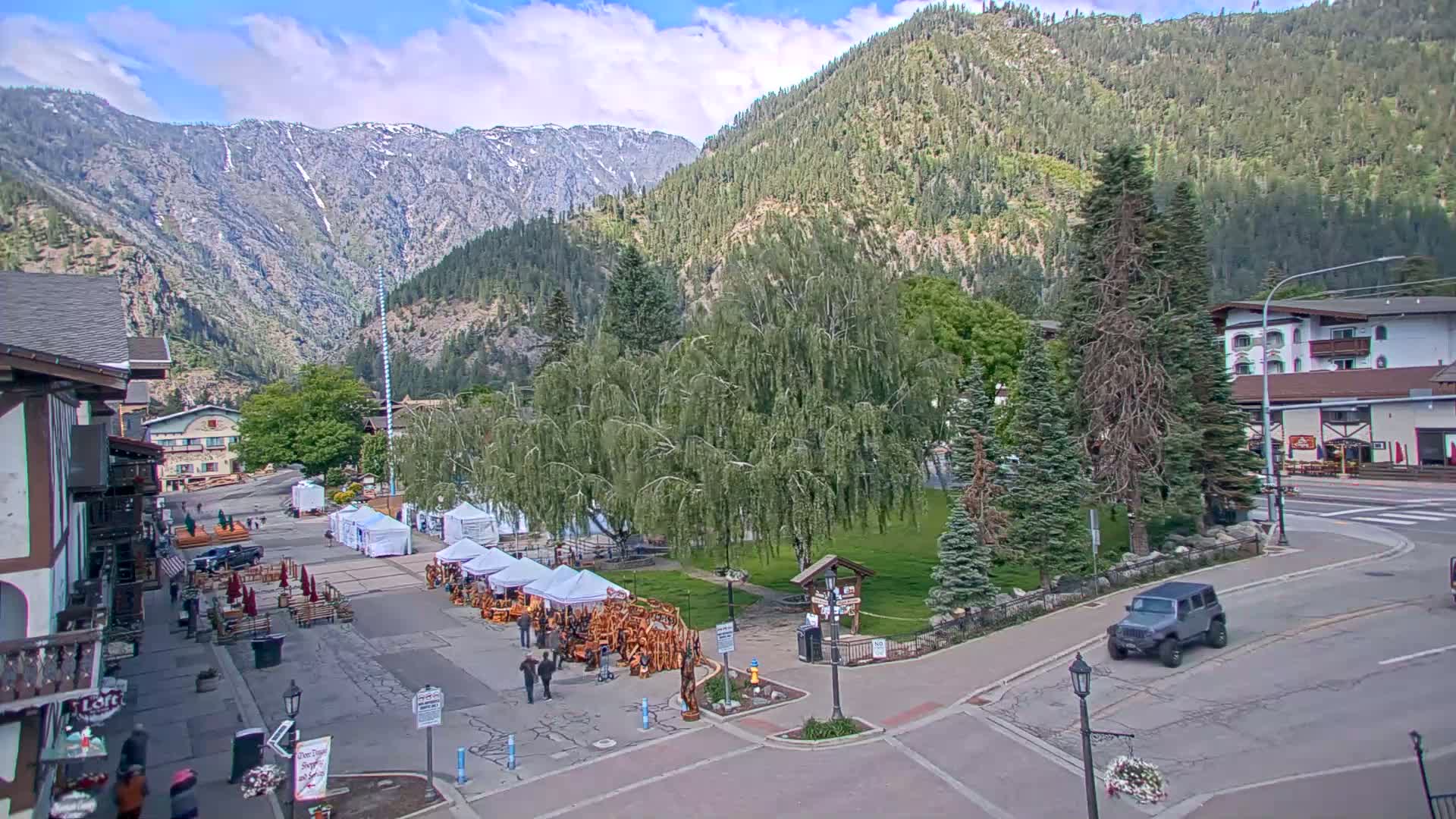 A mountain town square with several market stalls, surrounded by buildings and trees, under partly cloudy skies, with mountains in the background.