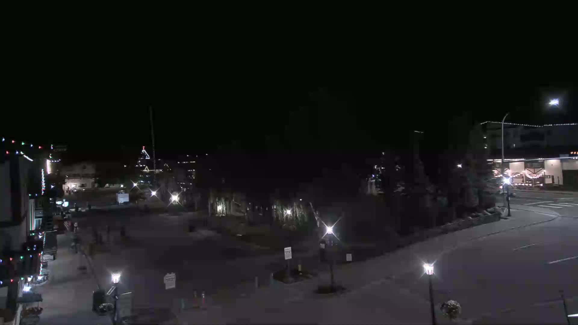 A nighttime, snow-covered town square is sparsely populated with a few people and buildings decorated with Christmas lights.