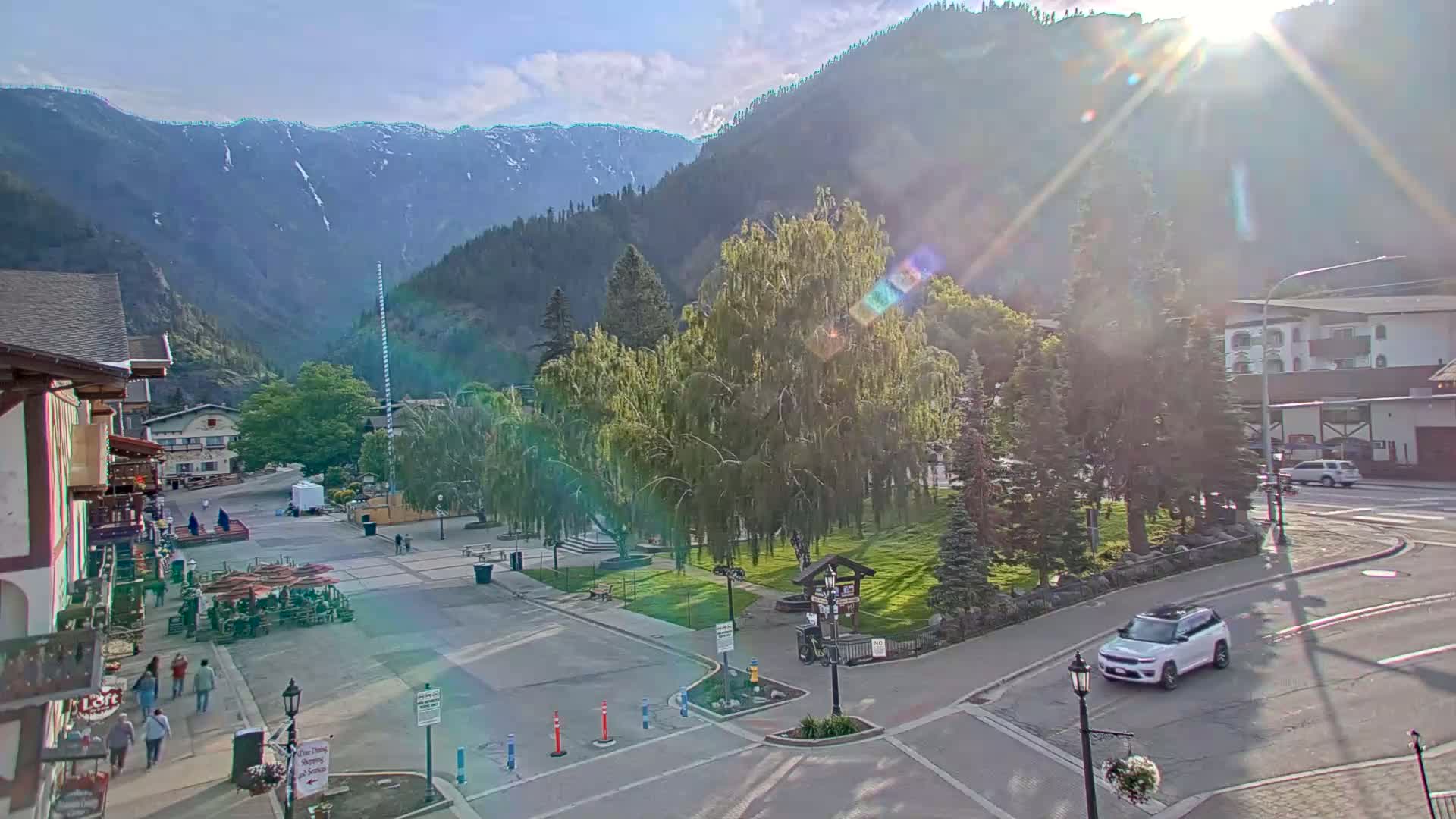 A sunny mountain town square with a grassy park in the center, surrounded by buildings and trees, with a few pedestrians and a car driving by.