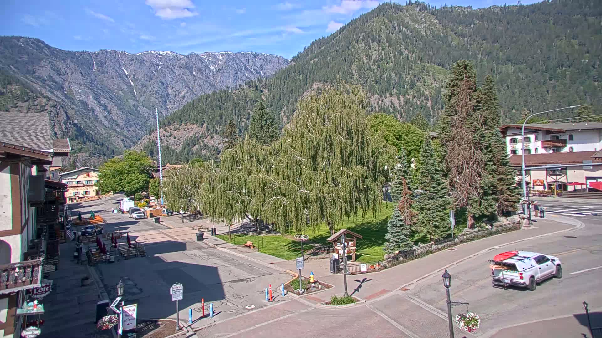 A sunny mountain town square with a weeping willow tree in the center, surrounded by buildings and a few cars, with mountains in the background.
