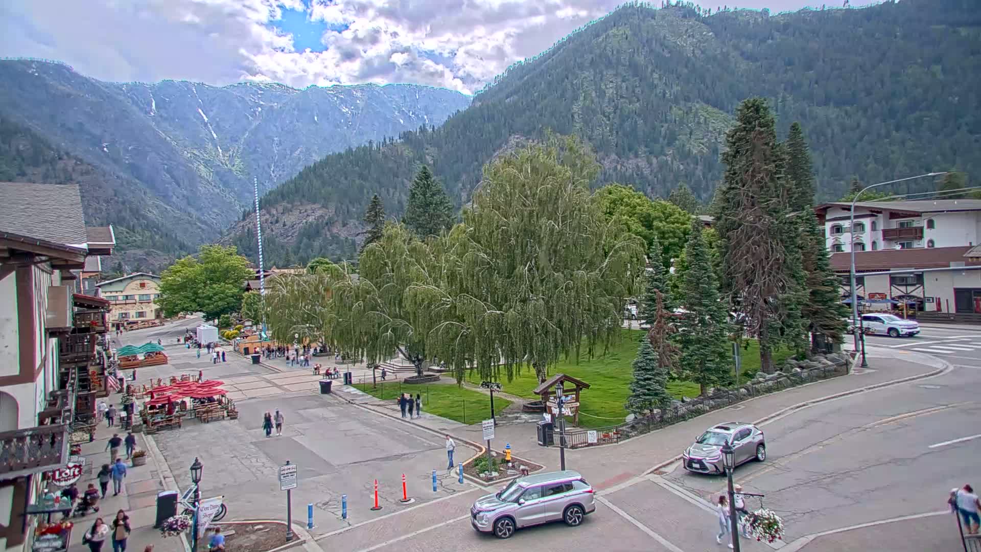 A partly cloudy day shows a mountain town square with people walking around, cars parked and driving, and a large weeping willow tree in the center of a grassy area, all set against a backdrop of mountains.