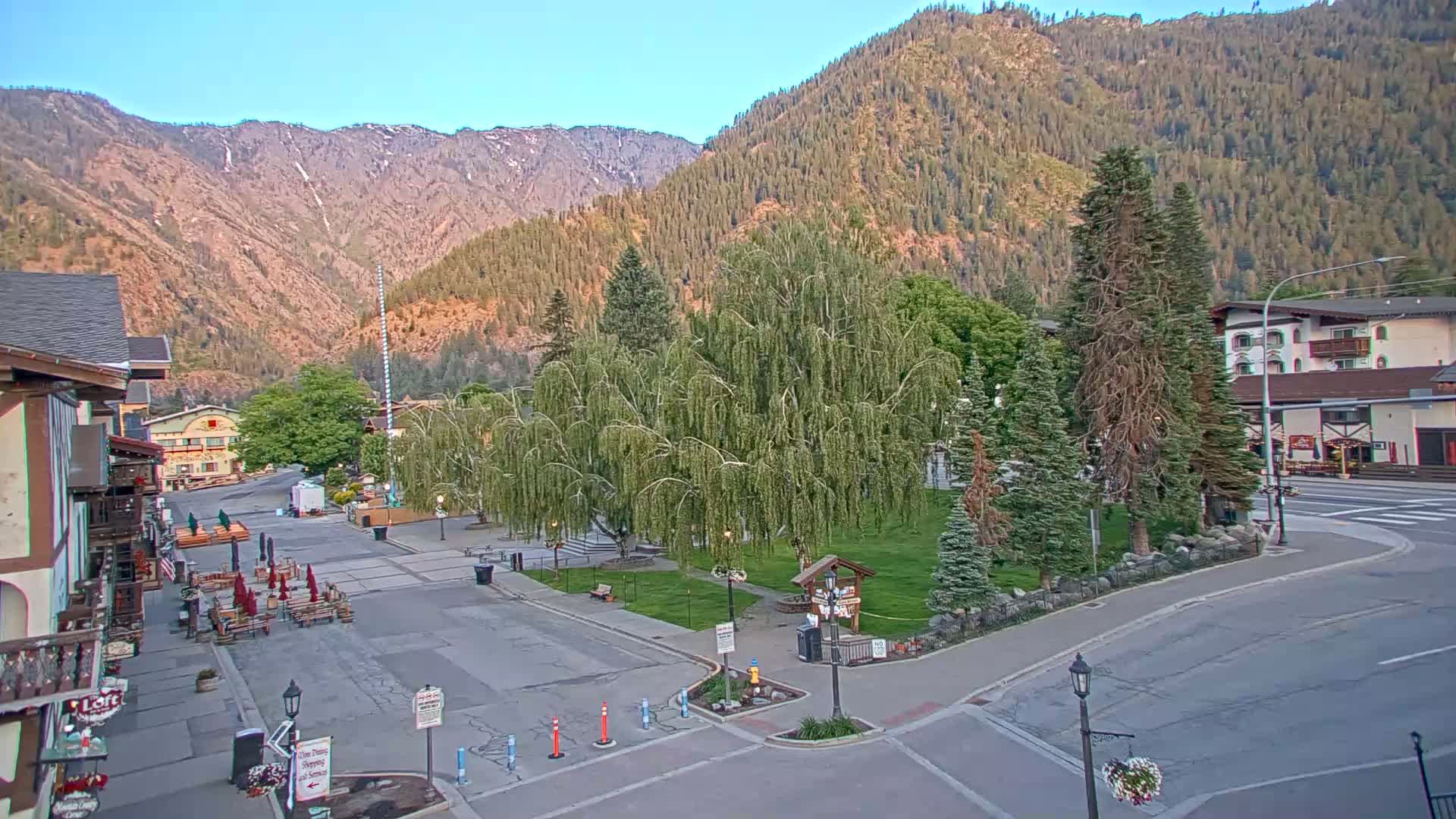 A mostly empty town square with a weeping willow tree in the center is situated at the base of a mountain range on a clear, sunny day.