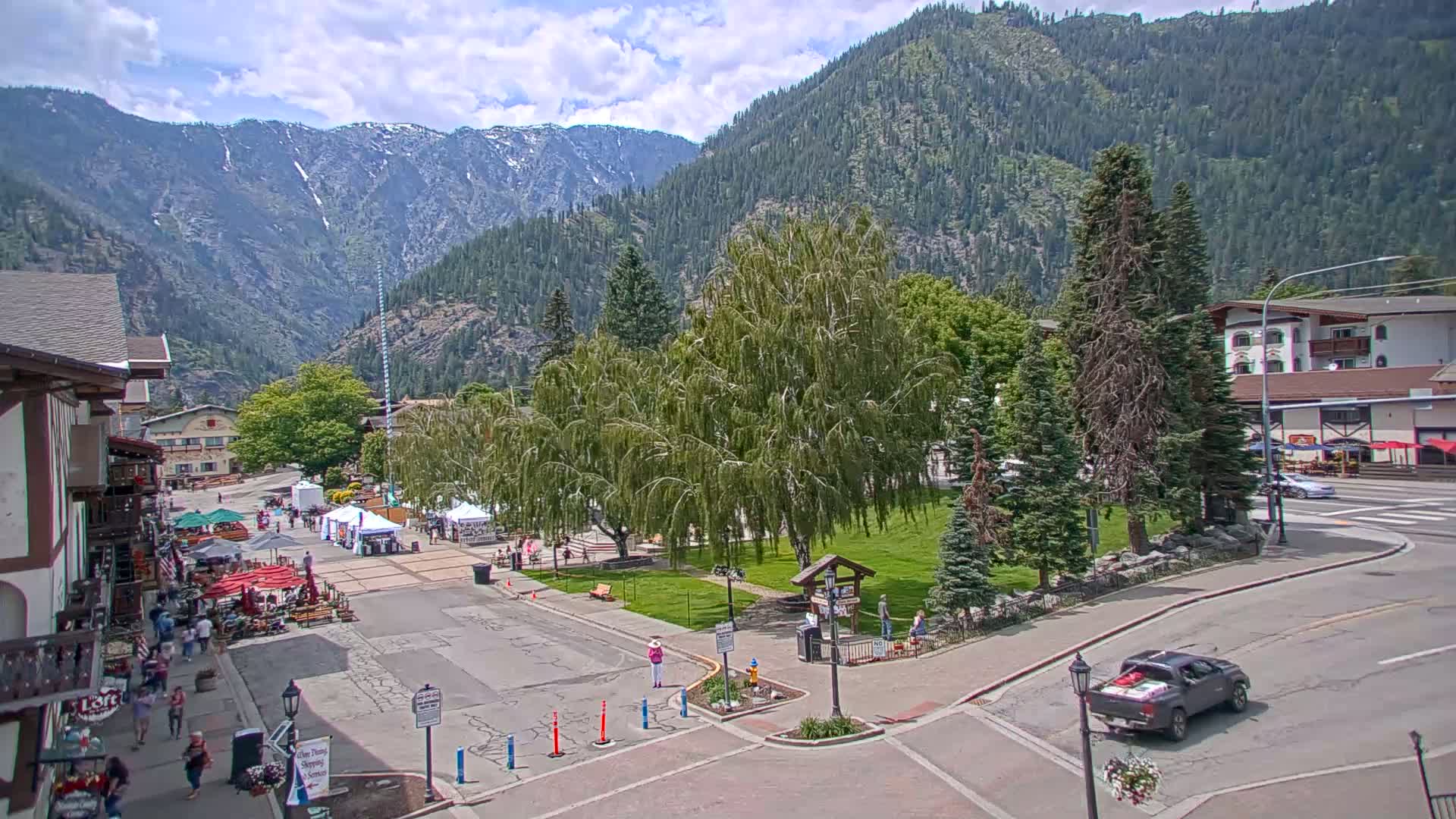 A partly cloudy day in a mountain town features a street scene with a small park, outdoor market stalls, and buildings, all set against a backdrop of snow-capped mountains.