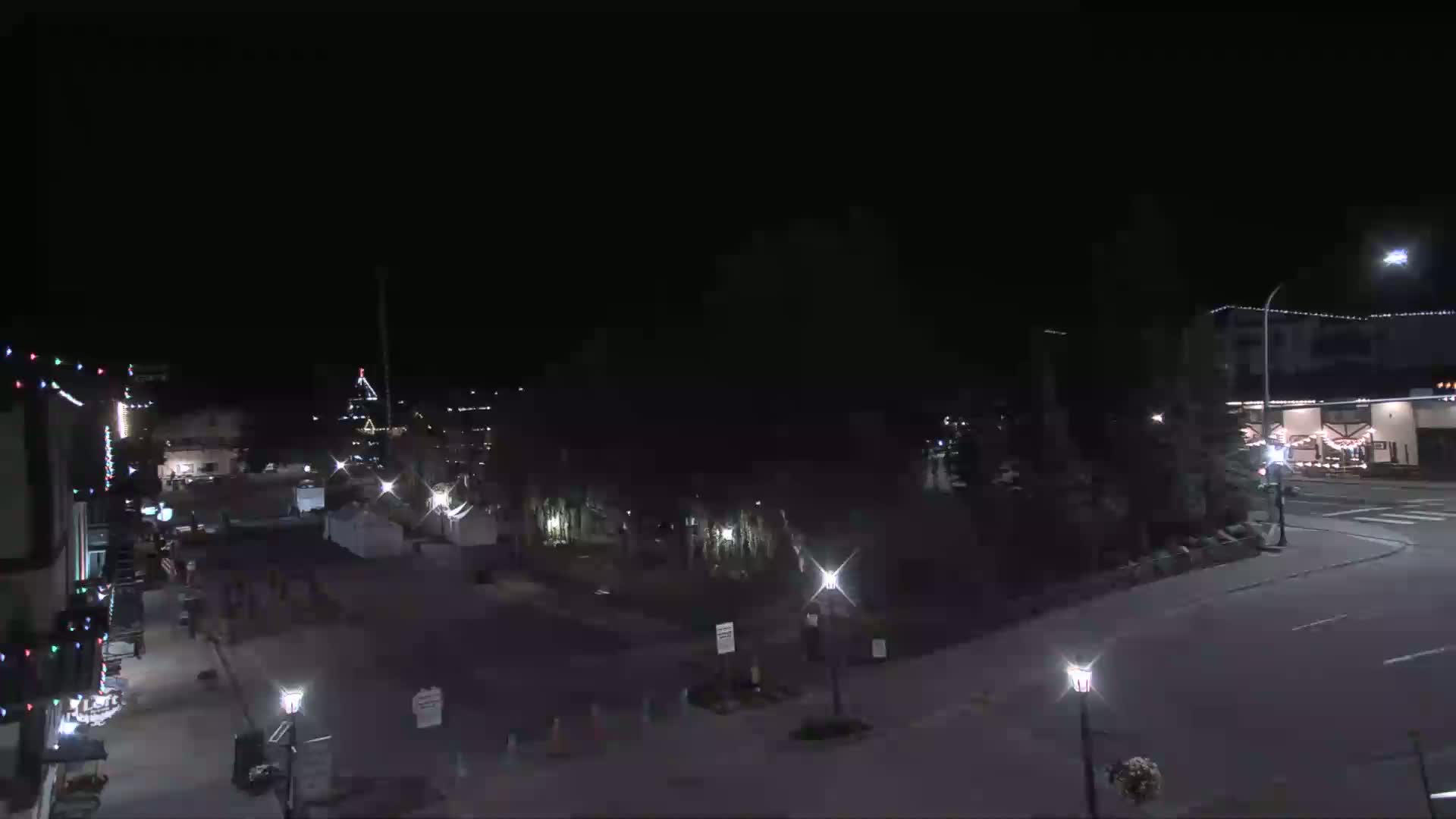 A nighttime, overhead view of a mostly empty town square with some buildings and streetlights illuminated.