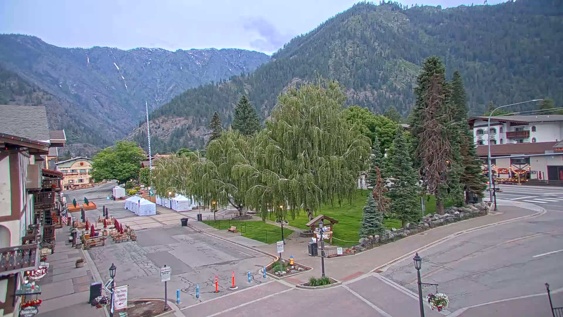 A mostly clear day reveals a town square nestled in a valley, surrounded by mountains, with several buildings and a large weeping willow tree.